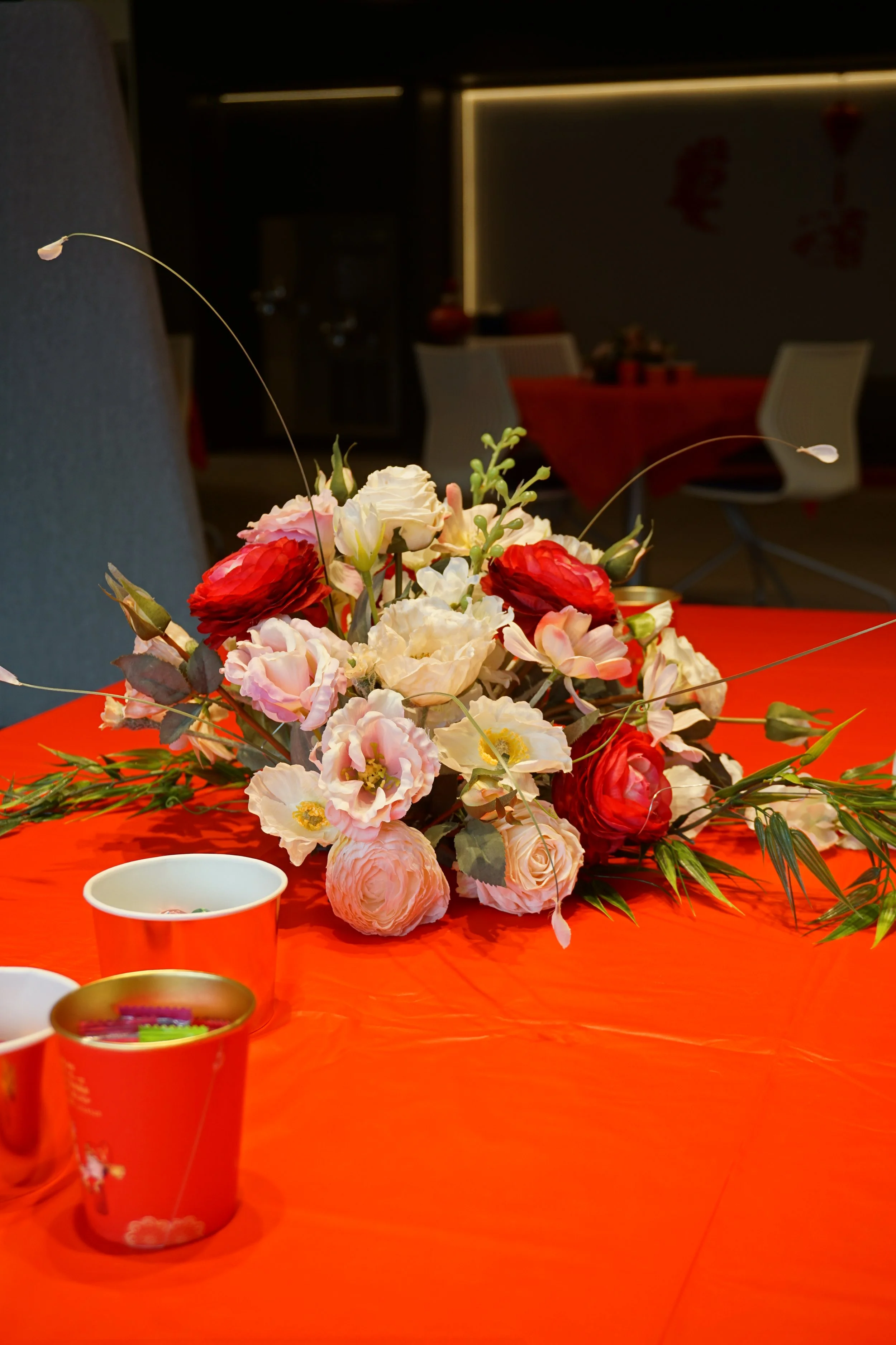A floral centerpiece with pink, white, and red flowers on a red tablecloth, with small cups nearby, in a dimly lit room.