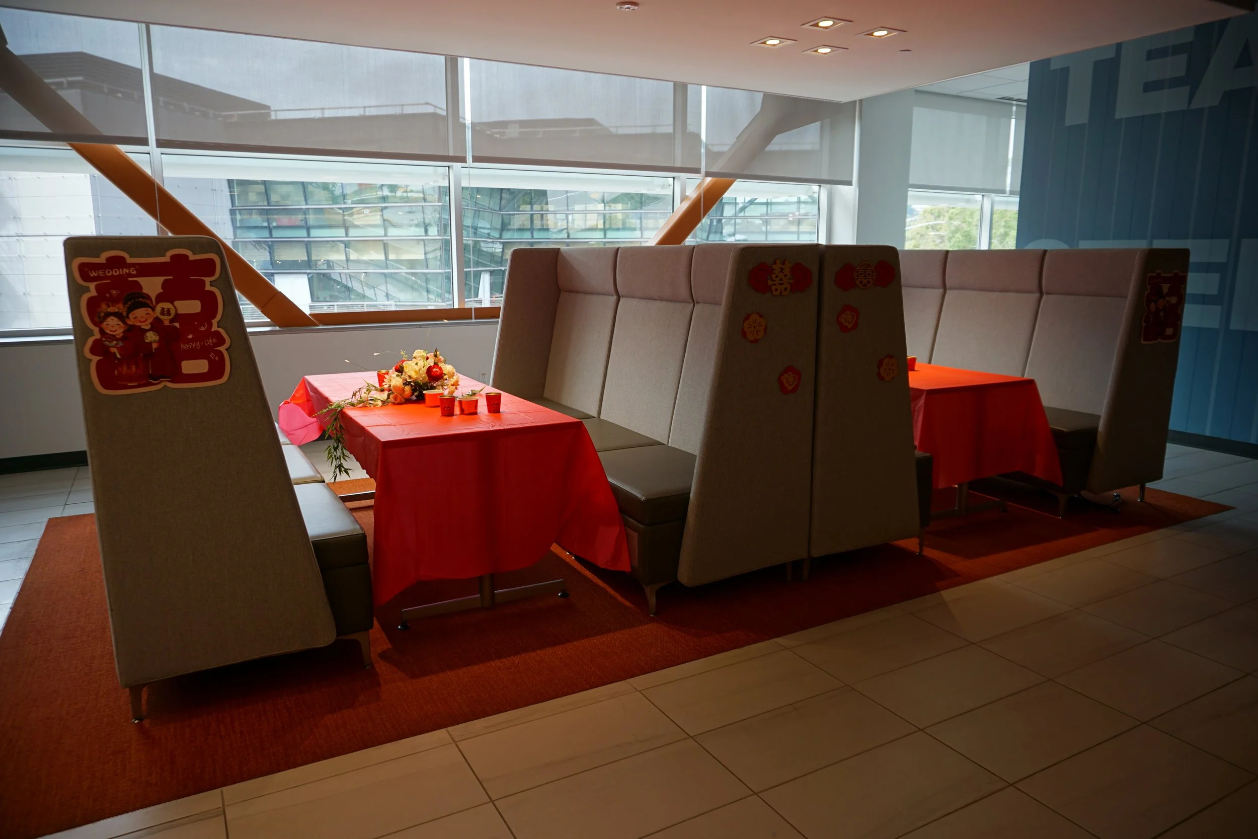 A decorated indoor dining area with two tables covered with red tablecloths, floral centerpieces, and small cups, enclosed by high-backed beige booths. Decorations featuring Chinese characters and themes suggest a celebration, possibly for a wedding 