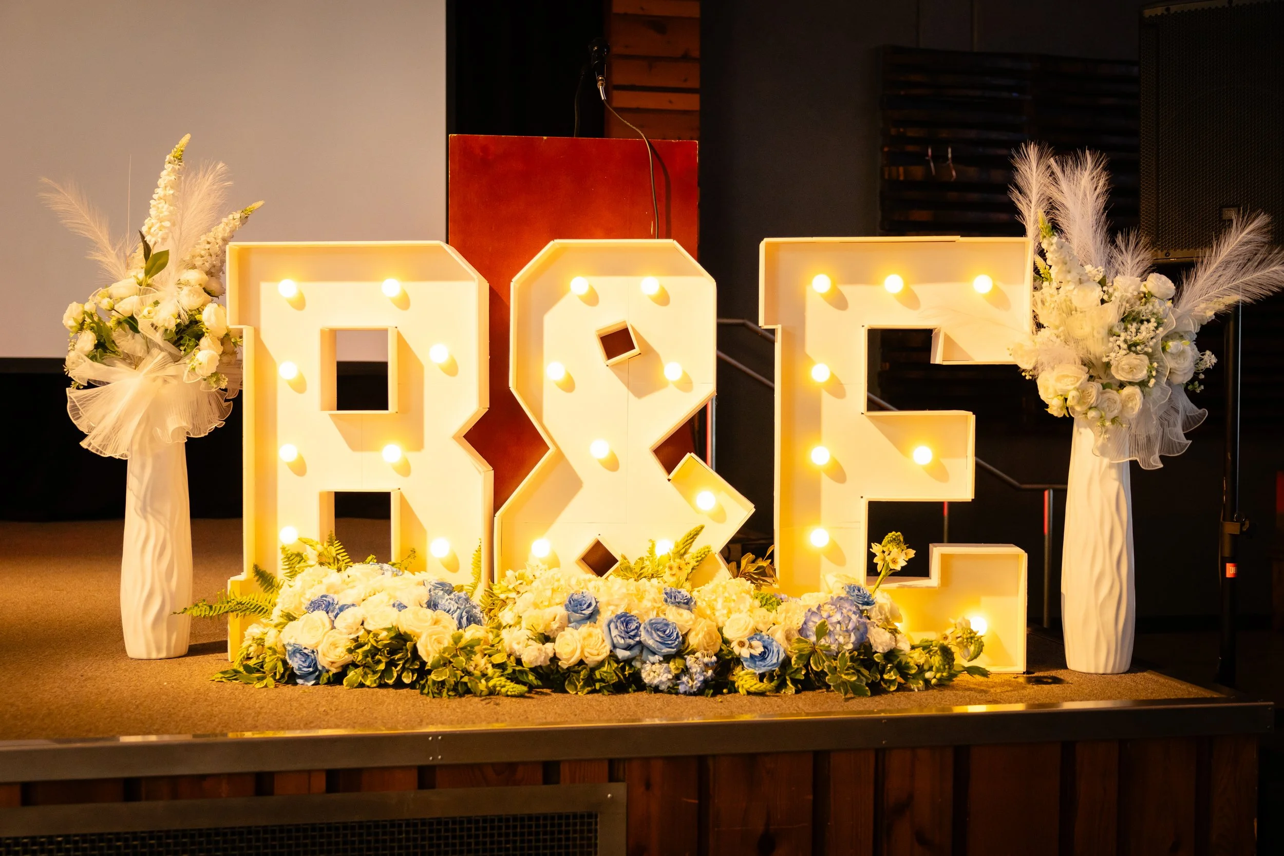 Decorative illuminated letters spelling 'LOVE' with white floral arrangements on either side and a bouquet of white and blue flowers at the base, set on a stage.
