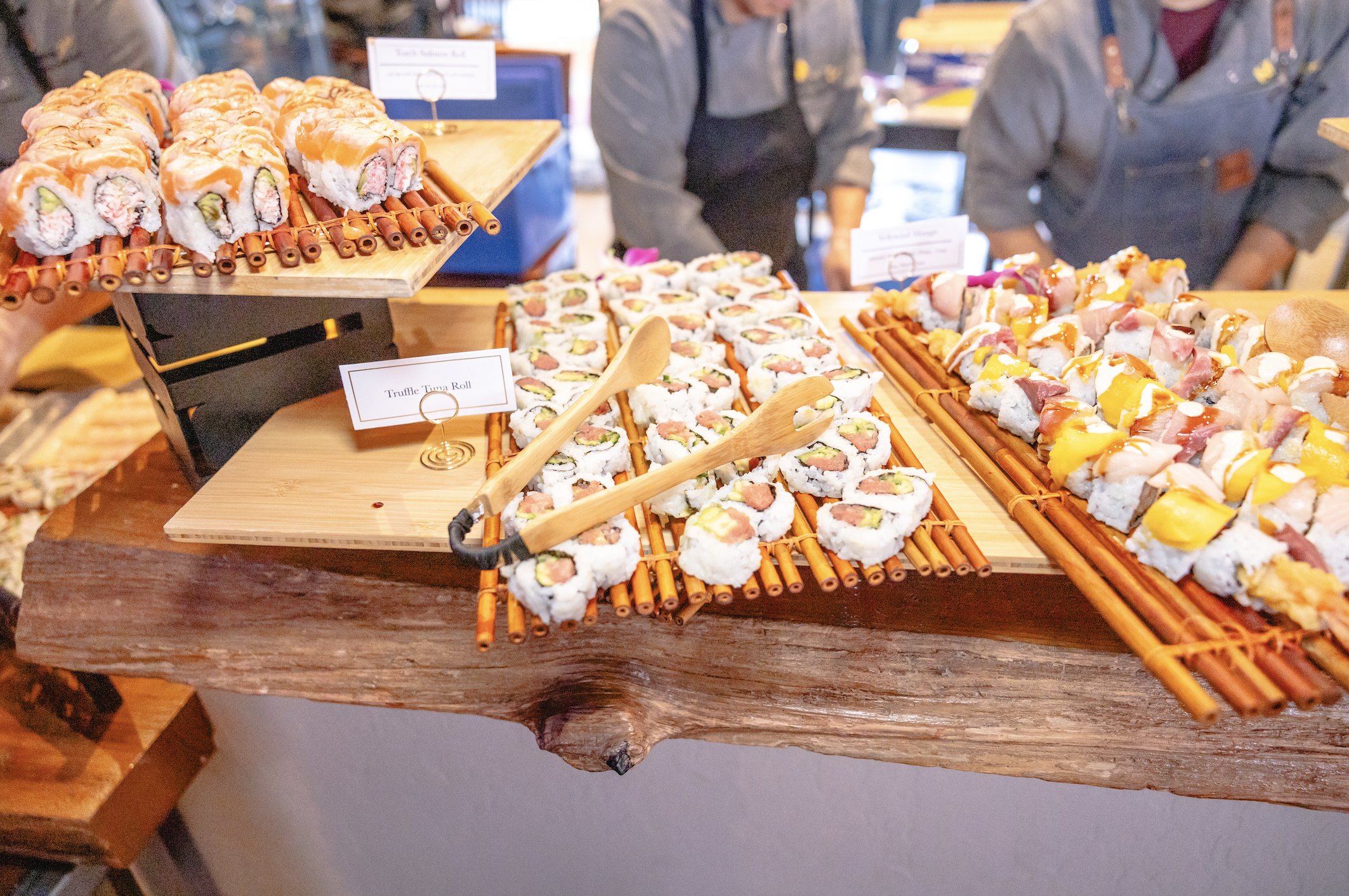 Assorted sushi rolls on bamboo mats, including tuna rolls and other seafood varieties, displayed on wooden surfaces in a sushi shop with people in the background.