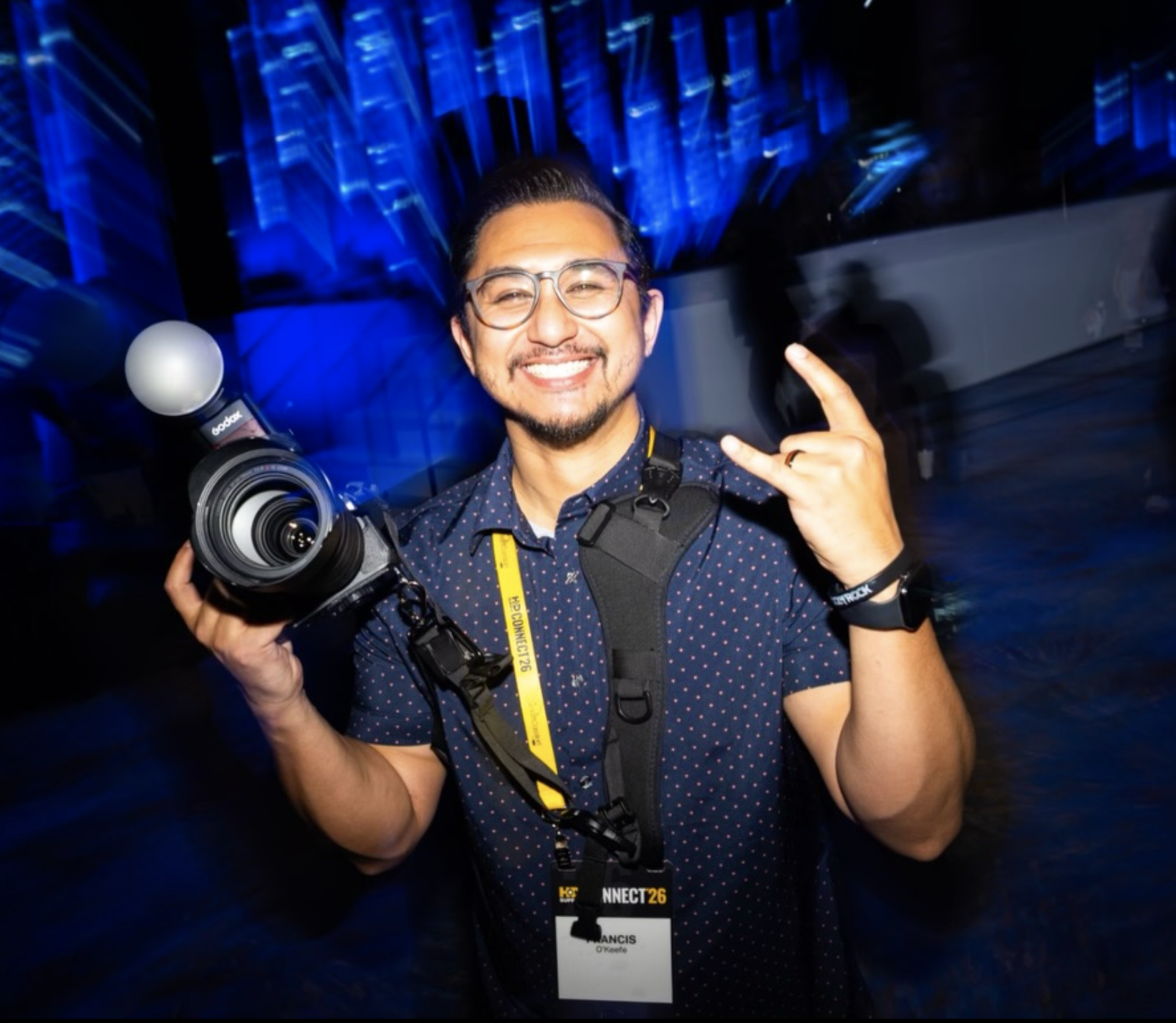 A smiling man with glasses holding a camera and making a rock hand gesture at a conference or event with blue lighting and motion effects.