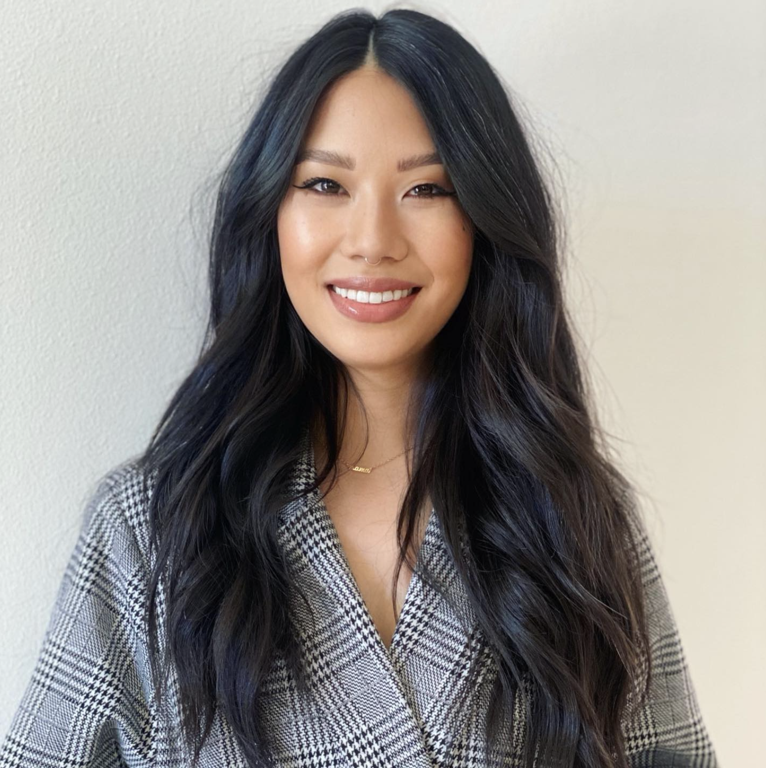 A woman with long black wavy hair smiling at the camera, wearing a gray plaid blazer, and a delicate gold necklace against a plain light-colored wall.