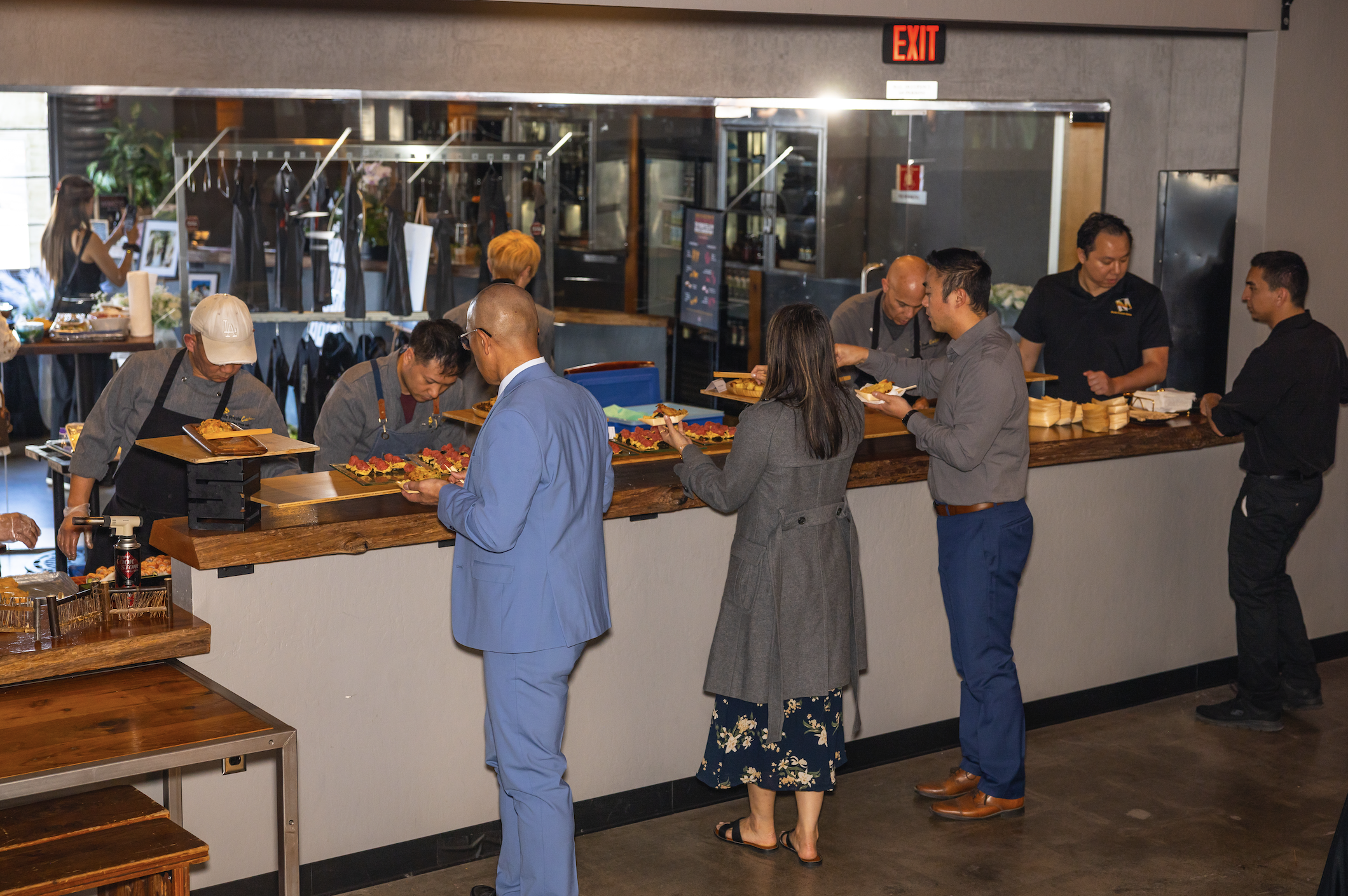 People standing in line ordering food at a restaurant counter with chefs and staff preparing meals.