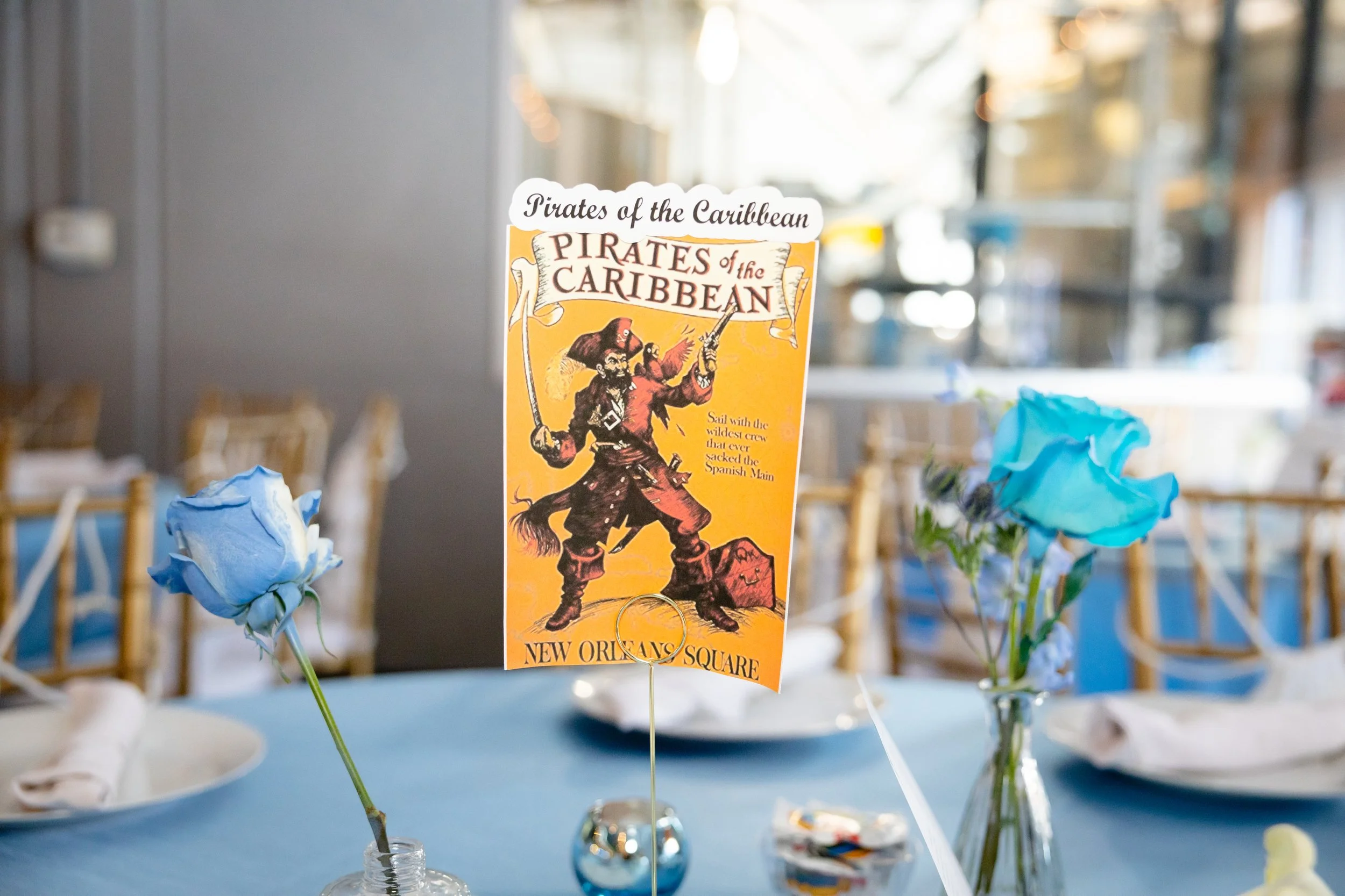 Table centerpiece with a card featuring a vintage pirate illustration, blue roses in small vases, and place settings at a party or event.