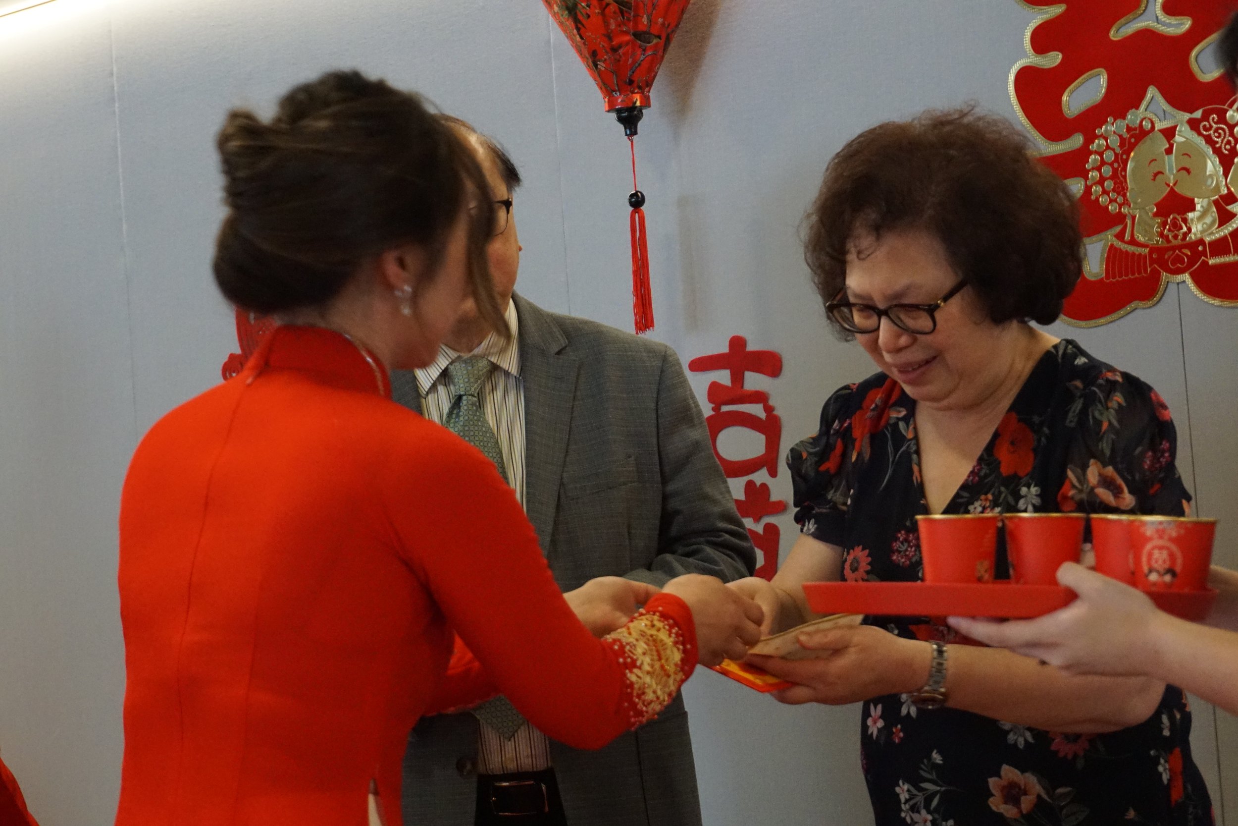 Women and man exchanging red envelopes during a Chinese celebration, with red and gold decorations in the background.