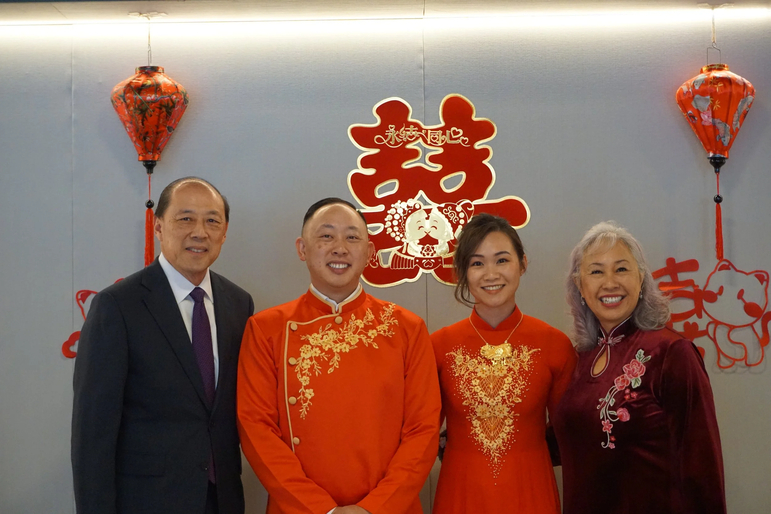 Four people standing together at a Chinese New Year celebration, with red lanterns and festive decorations behind them.