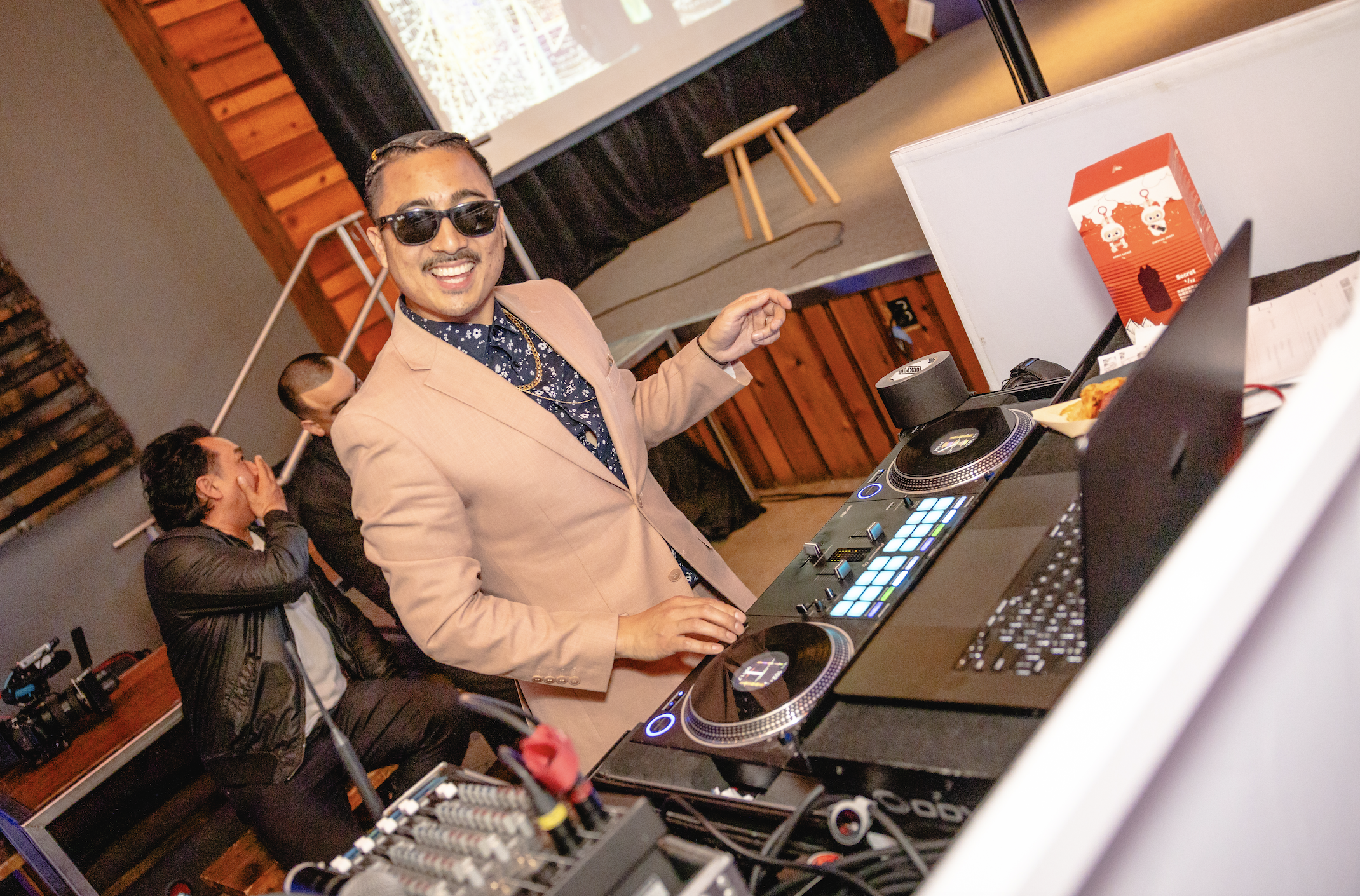DJ wearing sunglasses and a tan suit, smiling while using a DJ controller at a party or event.