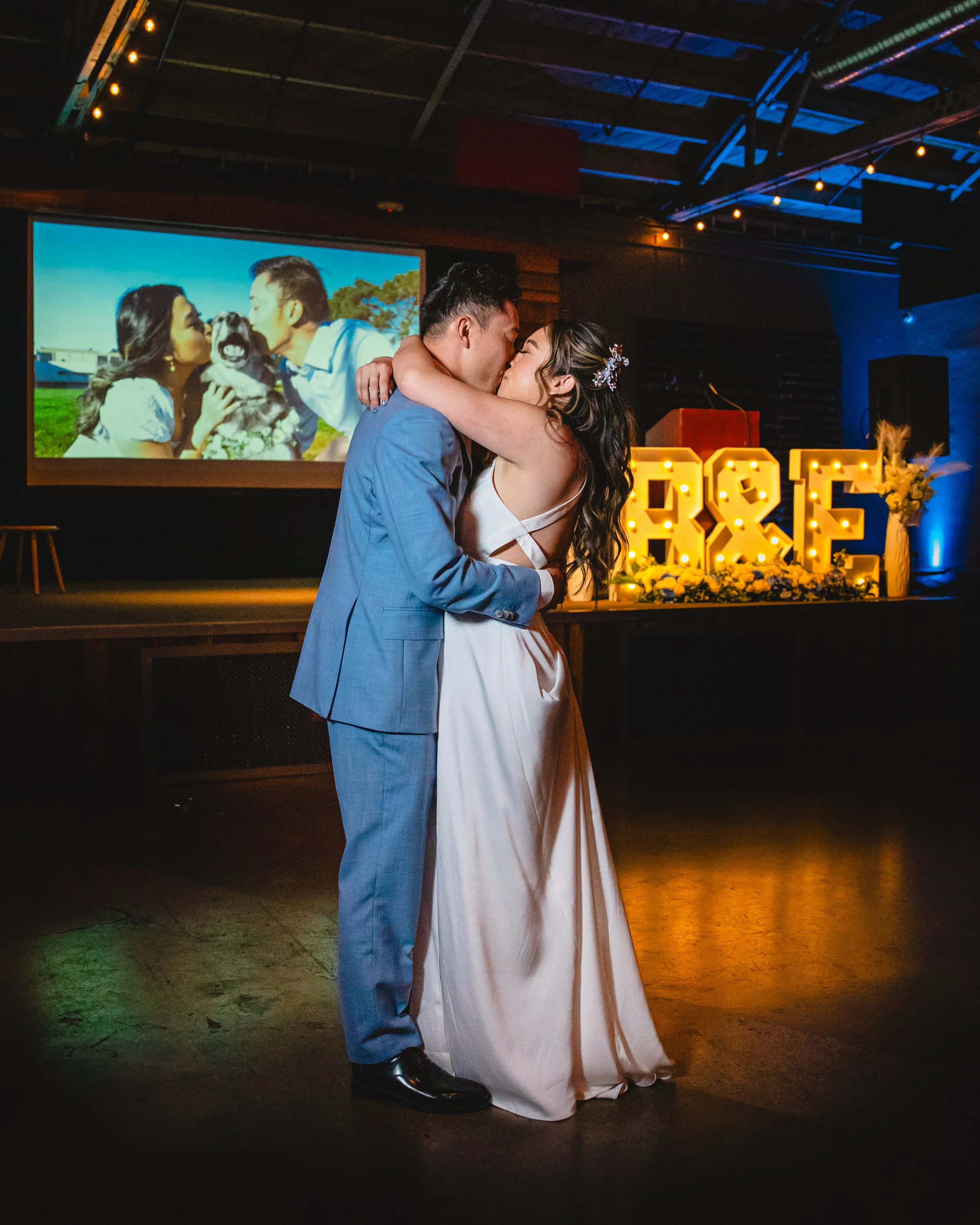 A newlywed couple sharing a kiss during their wedding reception with large illuminated letters 'A & E' and a large flower arrangement in the background. A slideshow image of the couple with their dog is projected on a screen behind them.