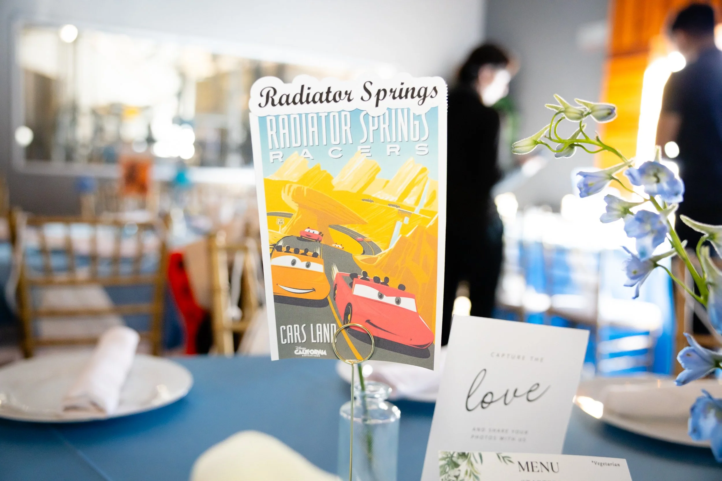 Table centerpiece with a colorful 'Radiator Springs Racers' poster from the Cars Land attraction, blue tablecloth, and a small white card that says 'love', with a flower arrangement on the right side.