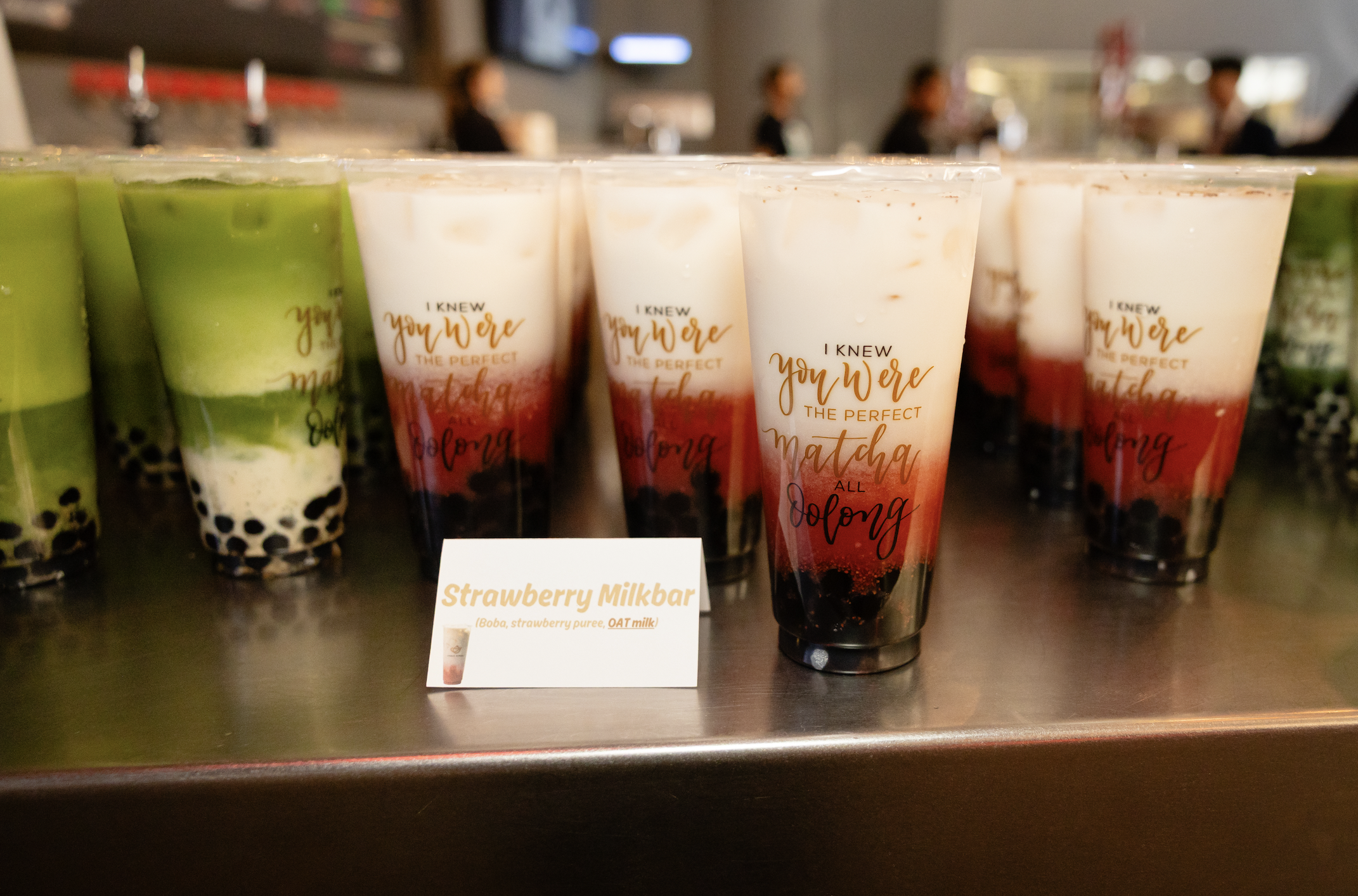 Rows of bubble tea drinks with layered colorful contents on a metal counter at a bubble tea shop, featuring a sign for a strawberry milkbar.