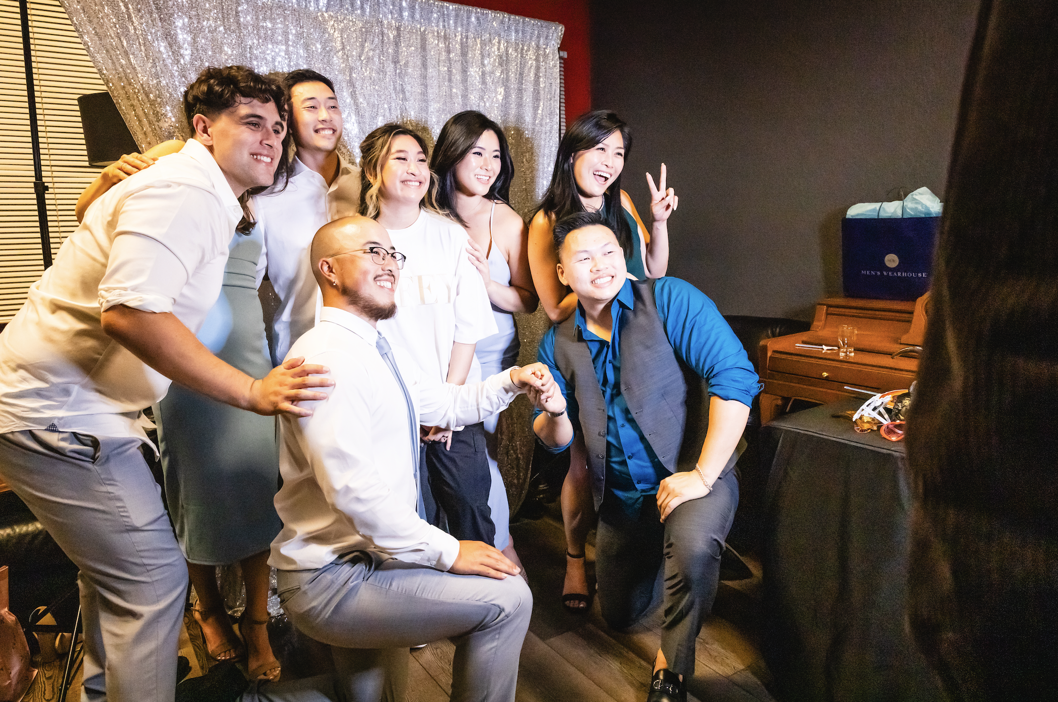 A group of seven friends posing for a photo in a dimly lit room, smiling and making peace signs, with a silver curtain backdrop and a piano in the corner.