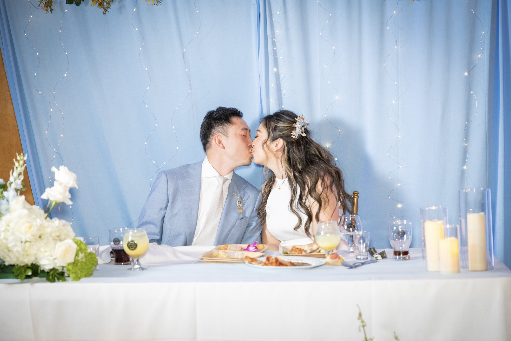 A couple kissing at a wedding reception table decorated with flowers, candles, and drinks against a blue backdrop with fairy lights.