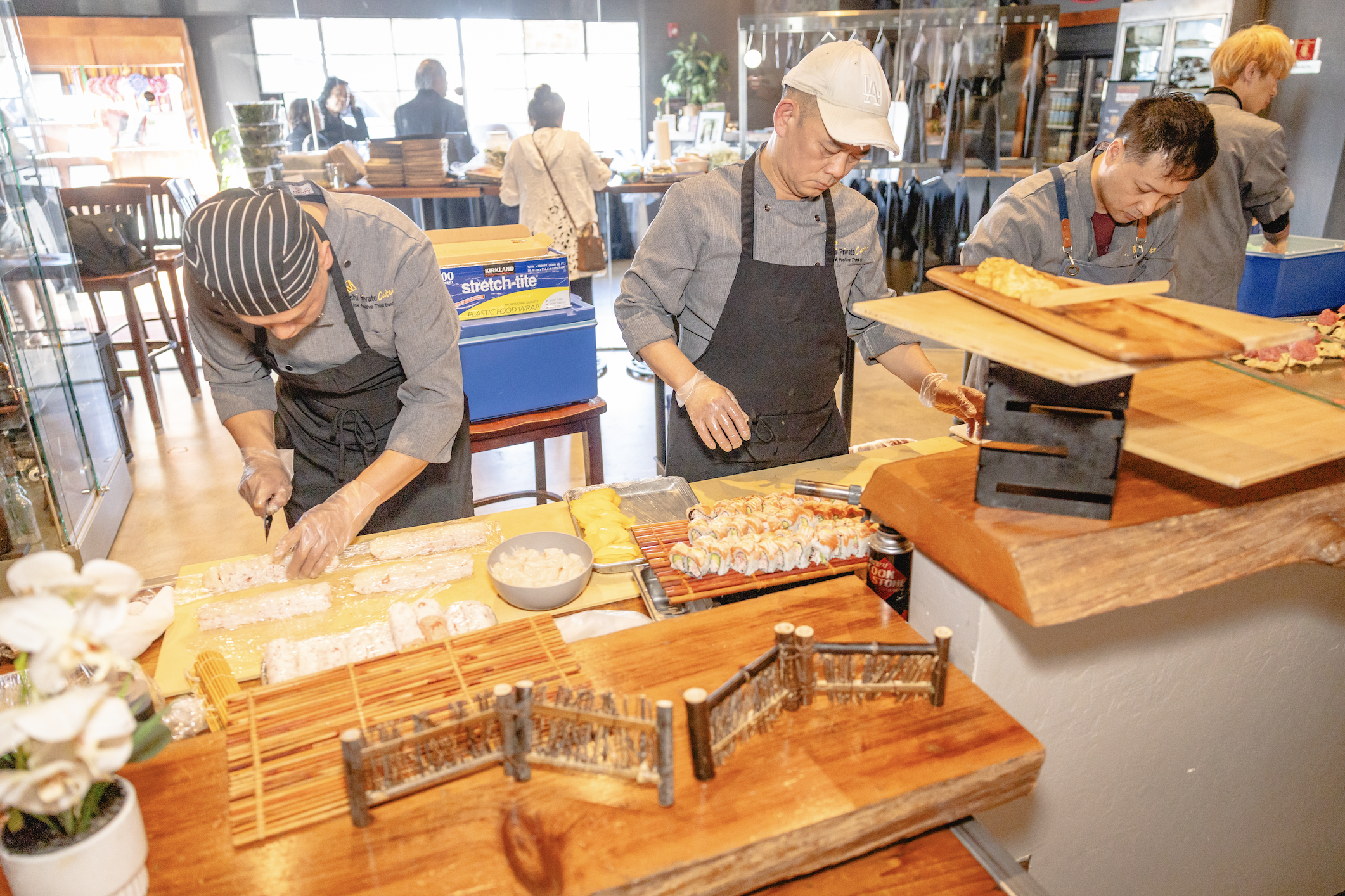 Three sushi chefs prepare sushi in a busy restaurant kitchen, with customers visible in the background.