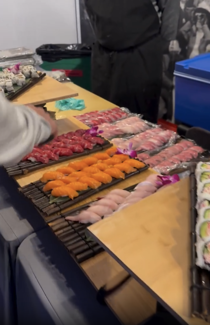 Sushi rolls and sashimi slices arranged on a wooden table at a sushi stand, with a person preparing sushi and a blue cooler in the background.