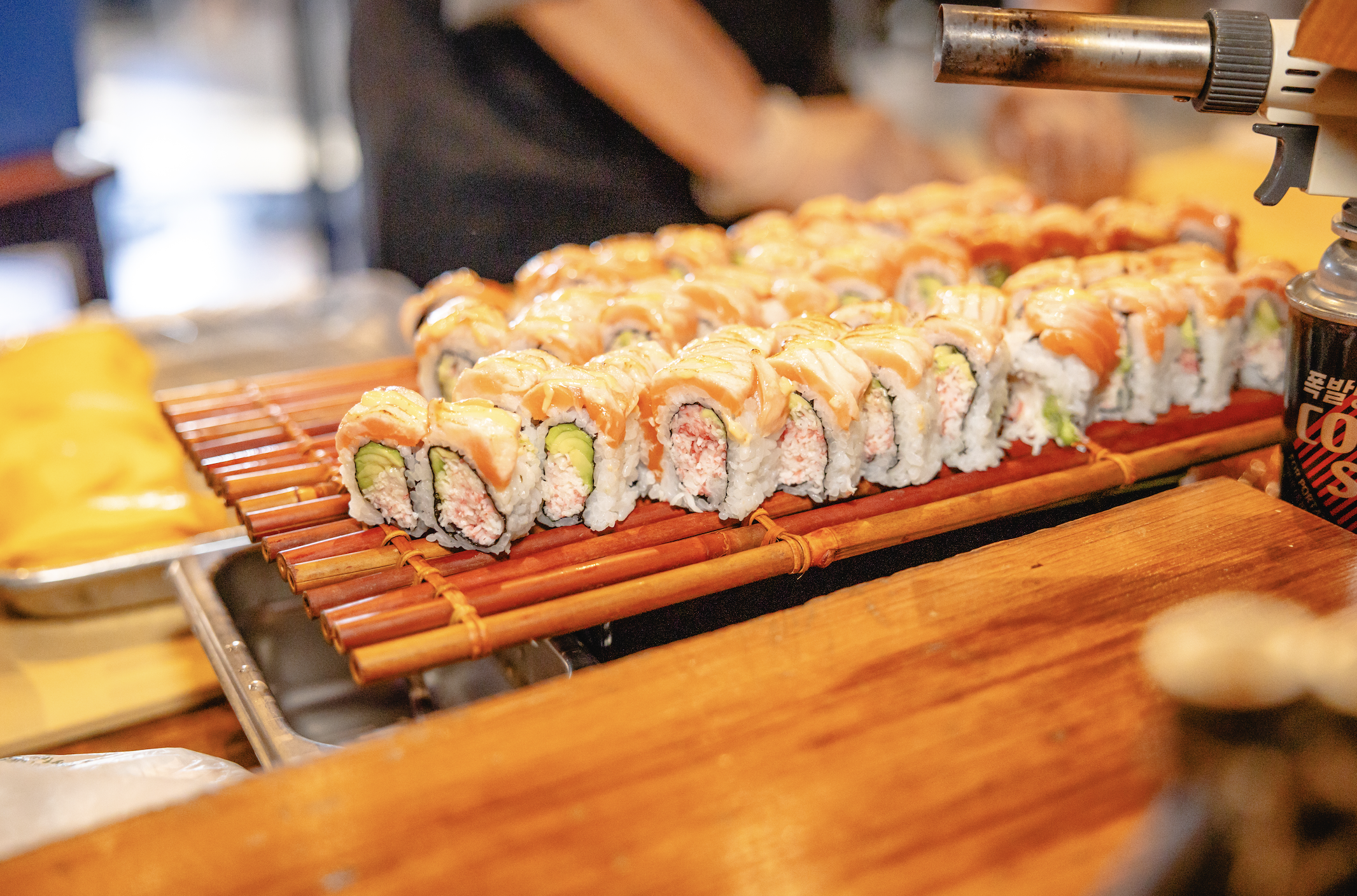 Close-up of sushi rolls with salmon, avocado, and crab, arranged on a bamboo mat at a food stall.