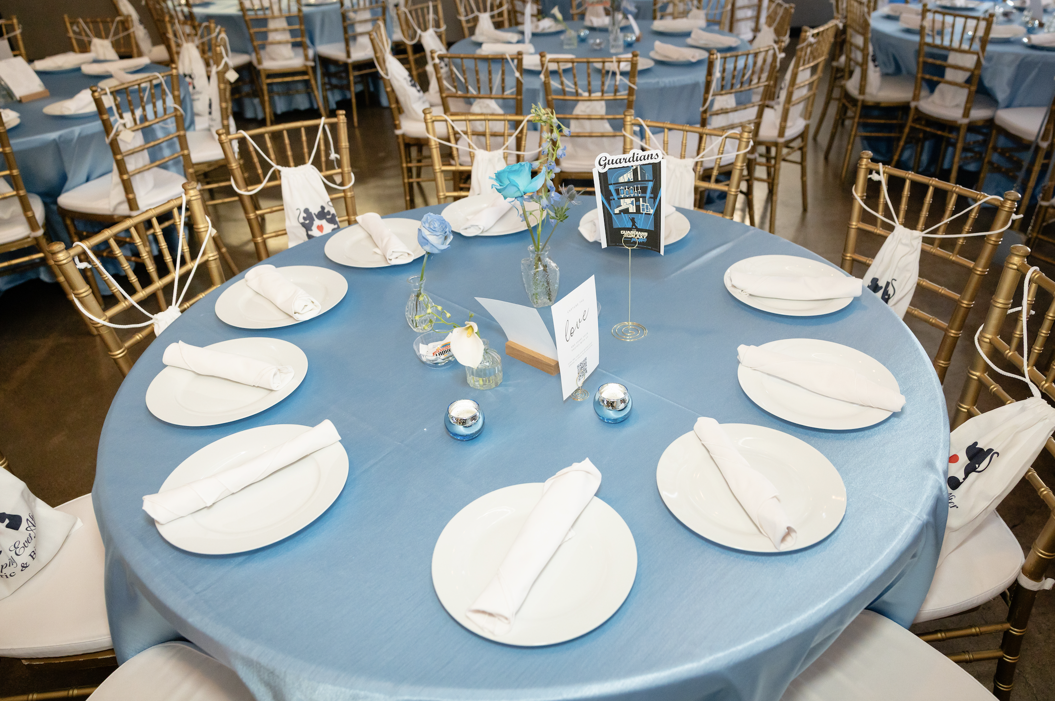 Round banquet table with a blue tablecloth, set with white plates, neatly rolled white napkins, small glass vases with blue flowers, and decorative items, surrounded by gold chairs with white cushions and white fabric decorations.