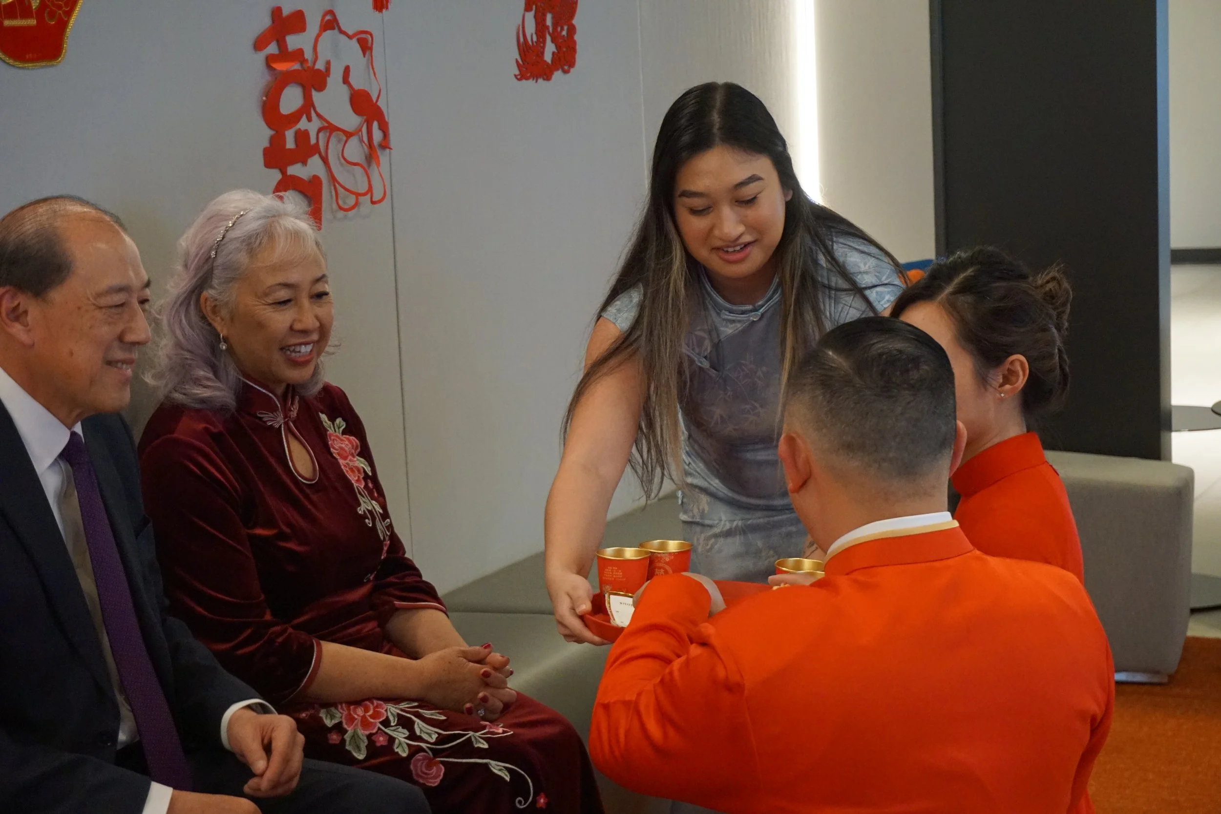 A woman is giving small cups to a man and woman dressed in traditional Chinese clothing, while three others sit nearby and watch, in a room decorated with red Chinese symbols and designs.
