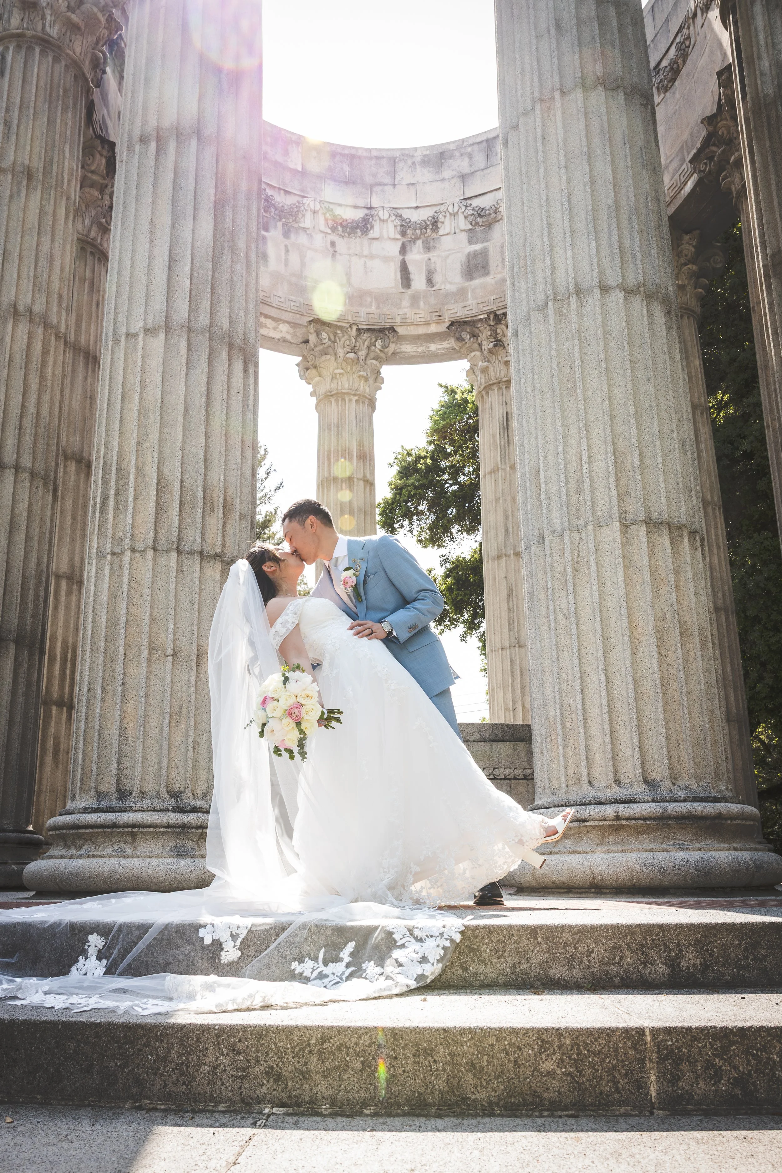 A bride and groom kiss at their wedding during the daytime, standing on steps in front of ancient Greek-style columns with sunlight shining down.