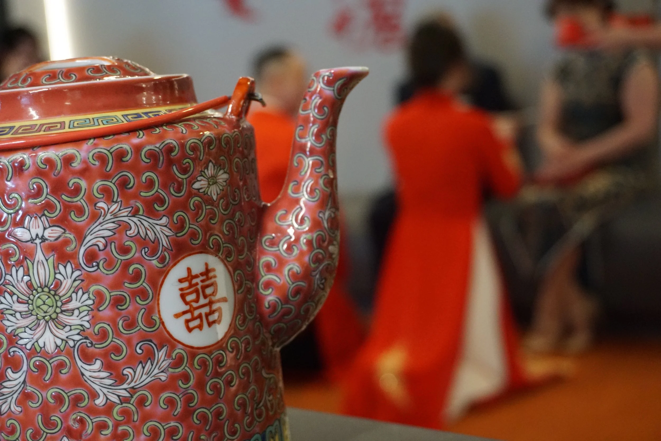 Close-up of a traditional ornate red teapot with Chinese characters and floral patterns, in focus, with people in red and black traditional clothing blurred in the background.