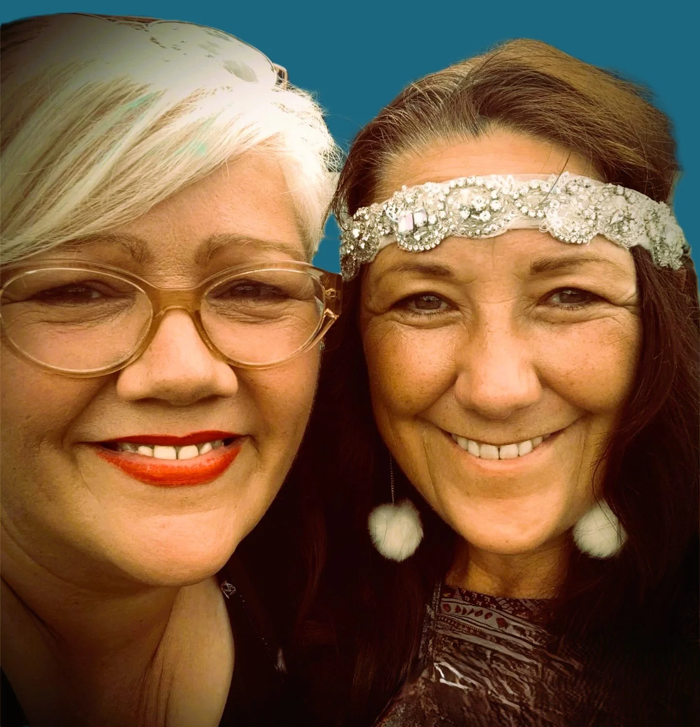 Close-up of two smiling women with styled hair and accessories, standing against a plain background.