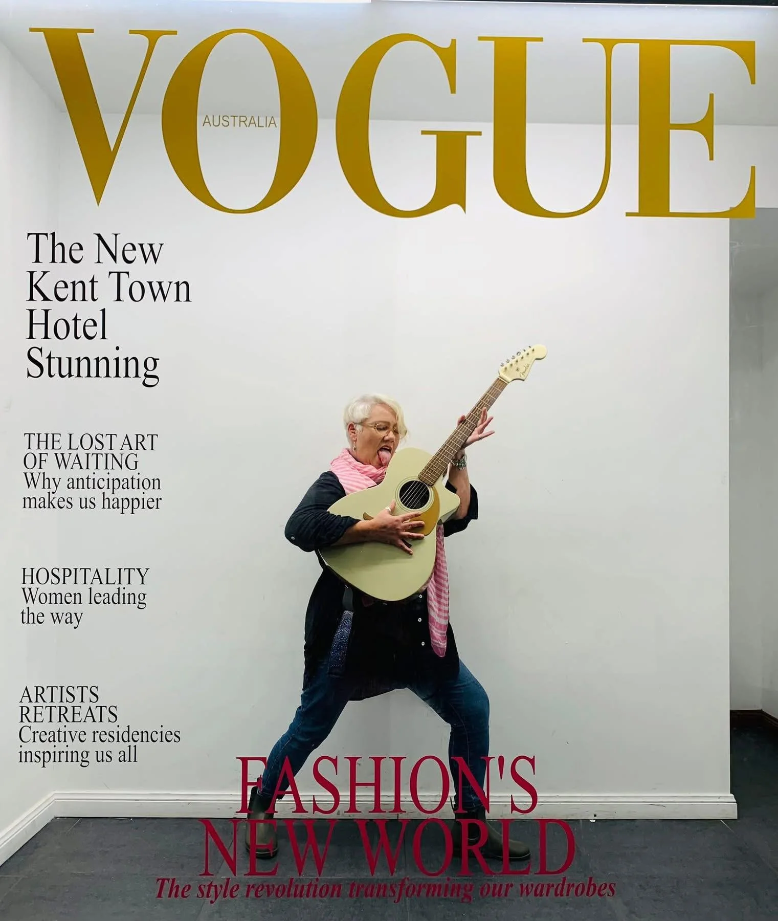 A woman with short gray hair playing an acoustic guitar and sticking out her tongue in front of a Vogue magazine cover. The cover features large gold text reading "VOGUE," with additional headlines about a hotel opening, art, hospitality, and fashion.