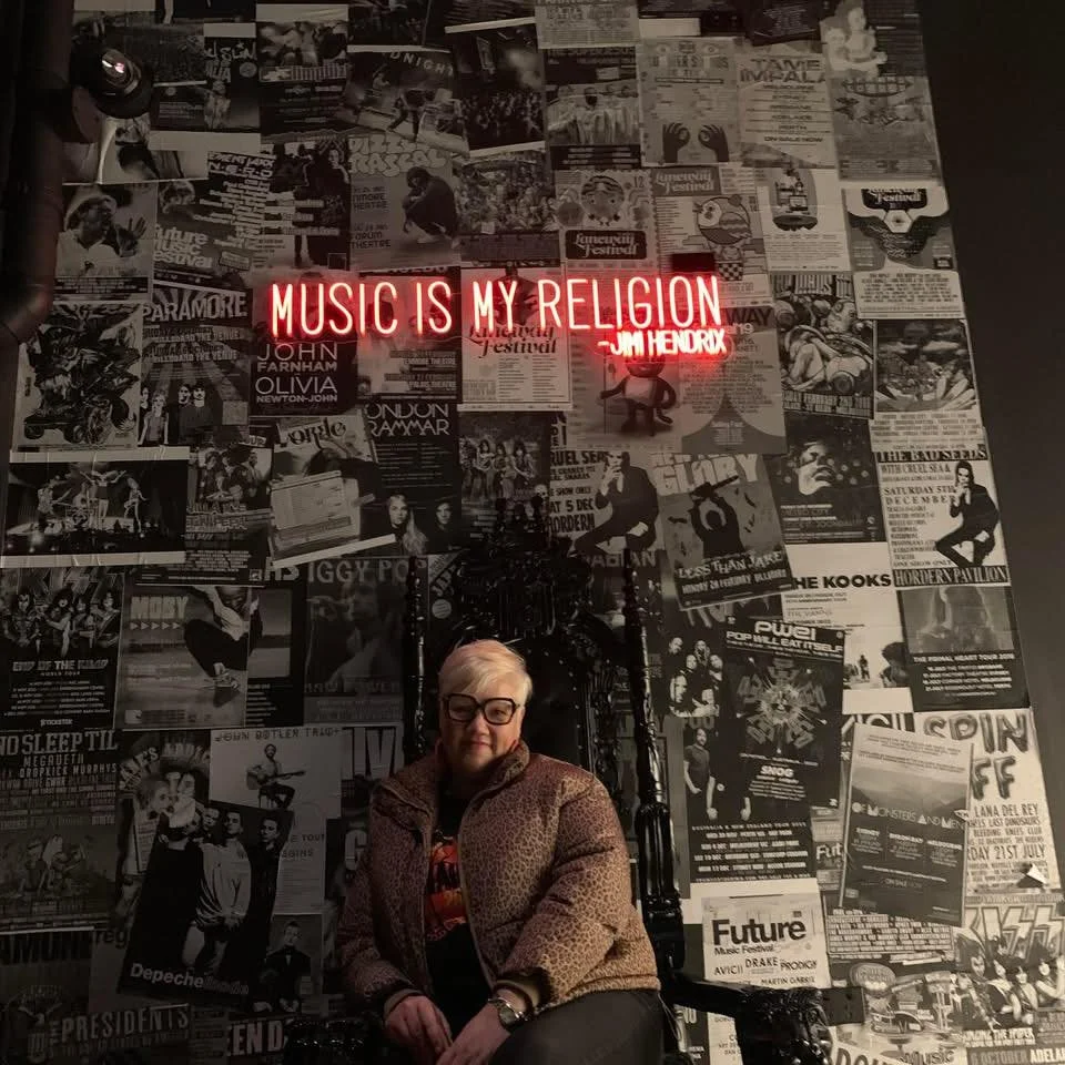 A woman with blond hair and glasses sitting on a black throne, with a background wall covered in black-and-white music and event posters. There is a red neon sign that says, "MUSIC IS MY RELIGION" with Jim Hendrix's name.