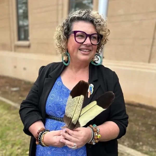 A woman with curly blonde hair and glasses smiling while holding a large bird feather in her hands, wearing a blue dress and several bracelets.