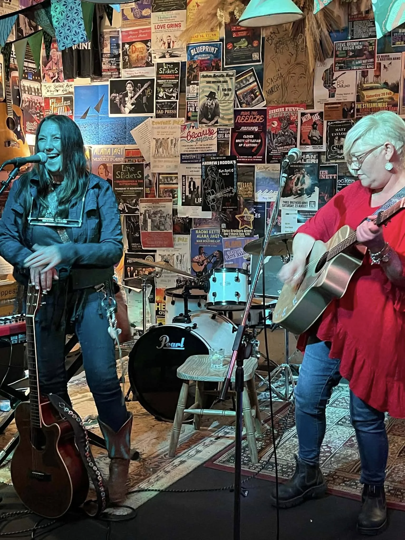 Two women performing with guitars on a small stage in a music venue decorated with posters and photos.