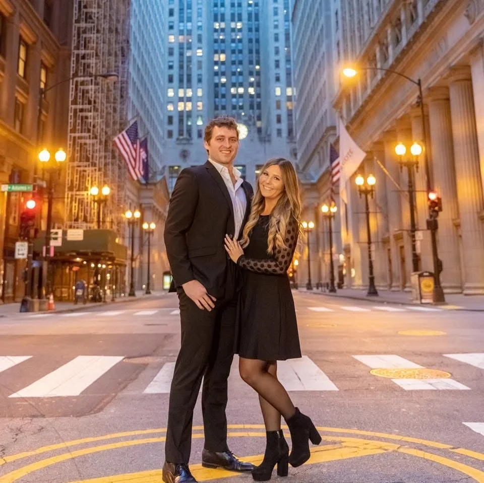 A man and woman standing close together on a city street at dusk, surrounded by tall buildings and illuminated streetlights, with American flags hanging from the buildings; the man is in a black suit, and the woman is in a black dress with sheer polka-dot sleeves.