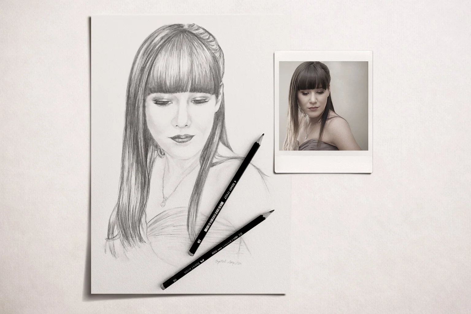 A pencil sketch of a woman with long hair and bangs, positioned next to a color photograph of the same woman with long, straight hair and bangs, both with a neutral expression. Two black pencils are placed on the sketch.
