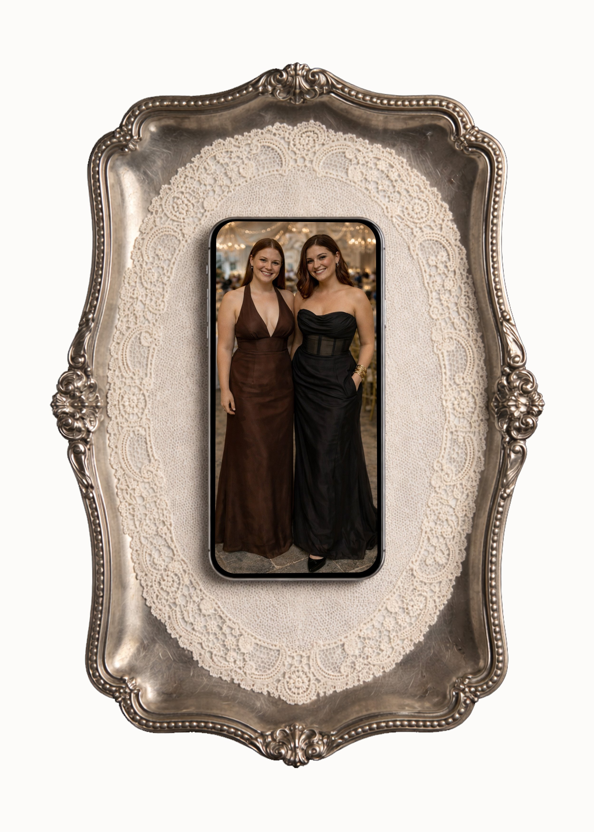Smartphone on ornate silver tray displaying an image of two women in evening gowns posing together at an event.