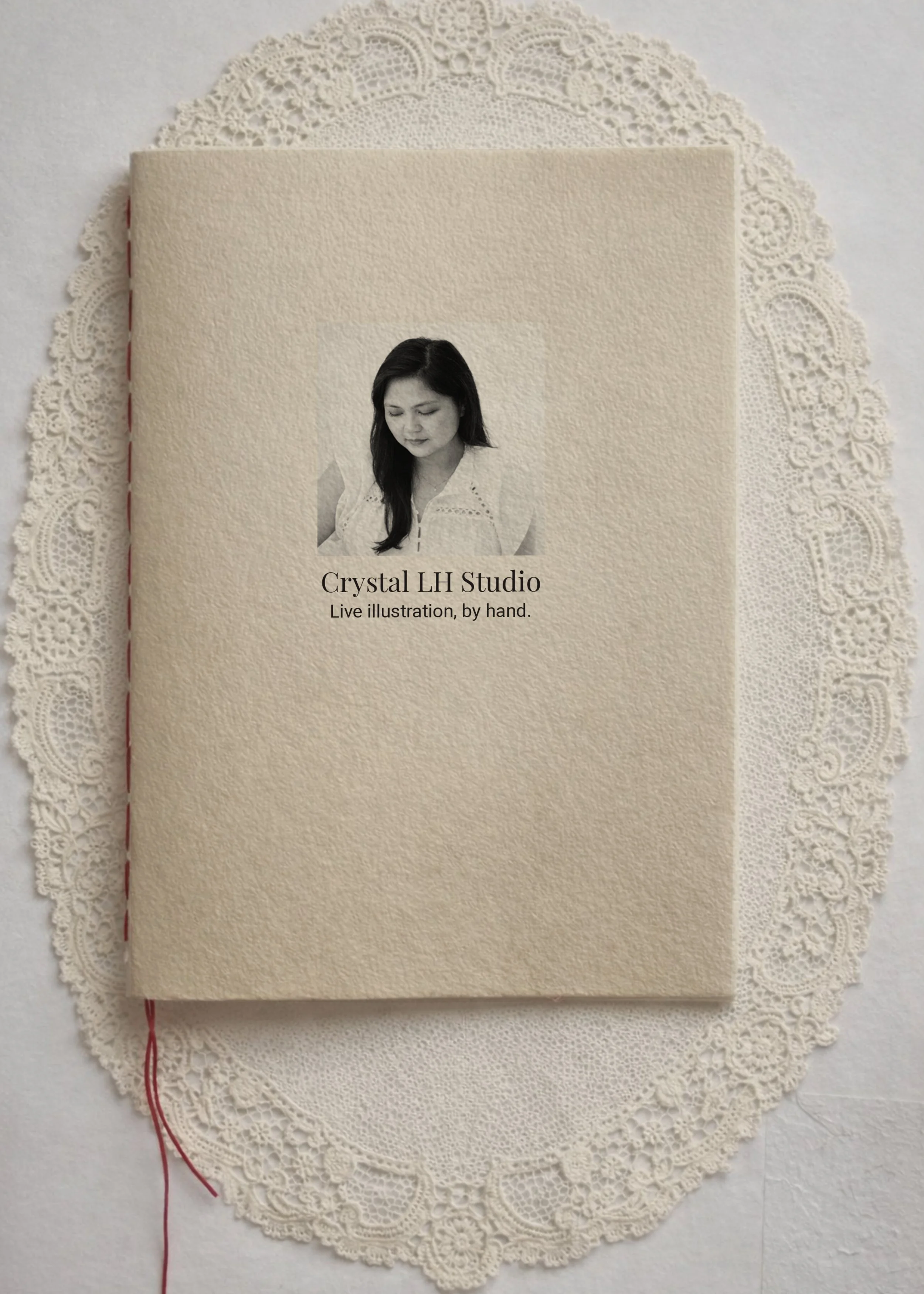 A closed cream-colored textured notebook with a small black-and-white photo of a woman on the cover. Text reads: 'Crystal LH Studio - Live illustration, by hand.' The notebook is placed on a decorative white lace doily.