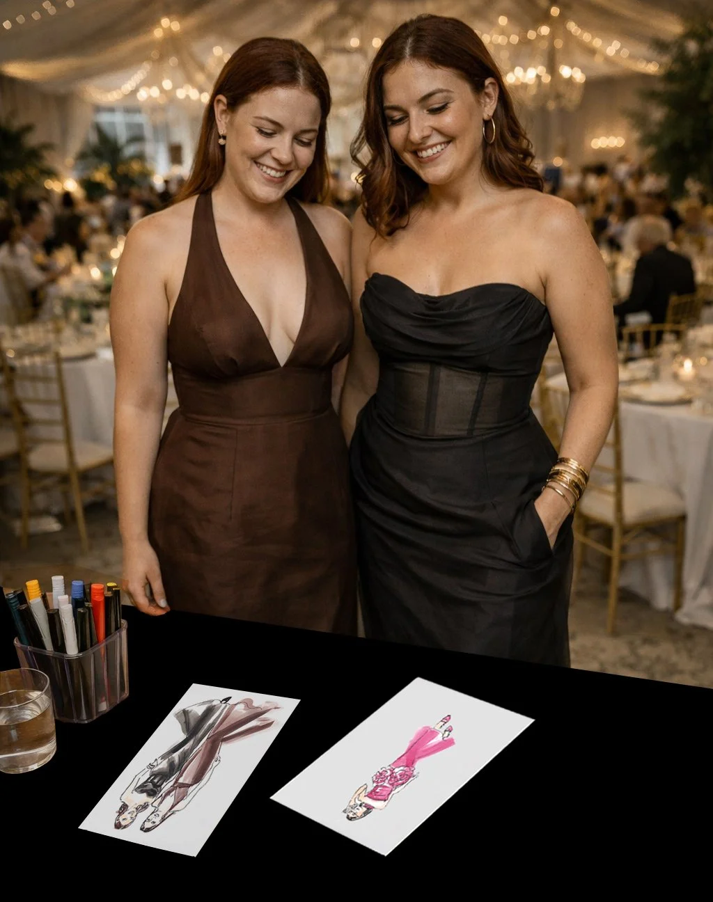 Two women smiling at a fashion illustration display at a formal event with decorated tables and warm lighting in the background.