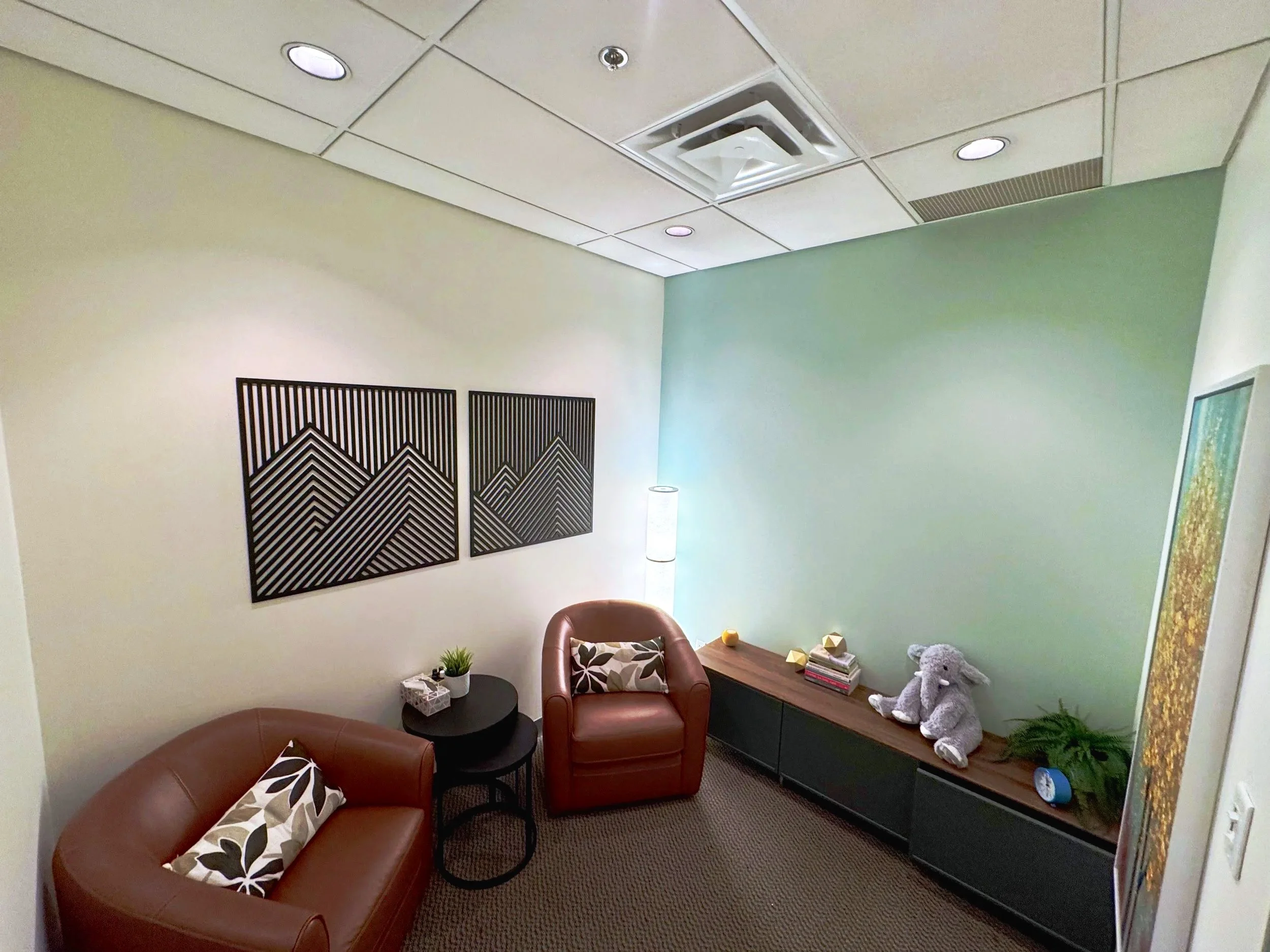 A cozy office with two brown leather armchairs, a black side table, wall art, and a wooden cabinet with decorative items, including a teddy bear and plants.