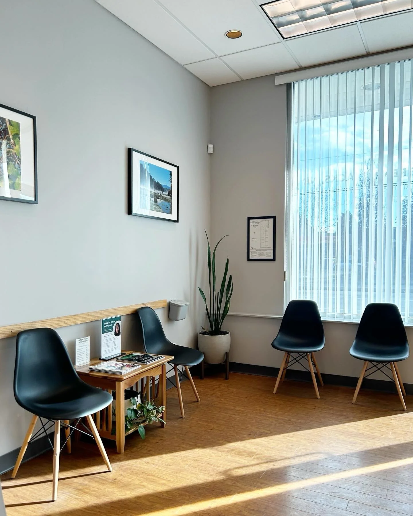 Warm & welcoming waiting room with four black chairs, a small wooden table with magazines, potted plant, framed pictures on the wall, large window with vertical blinds, and wooden flooring.