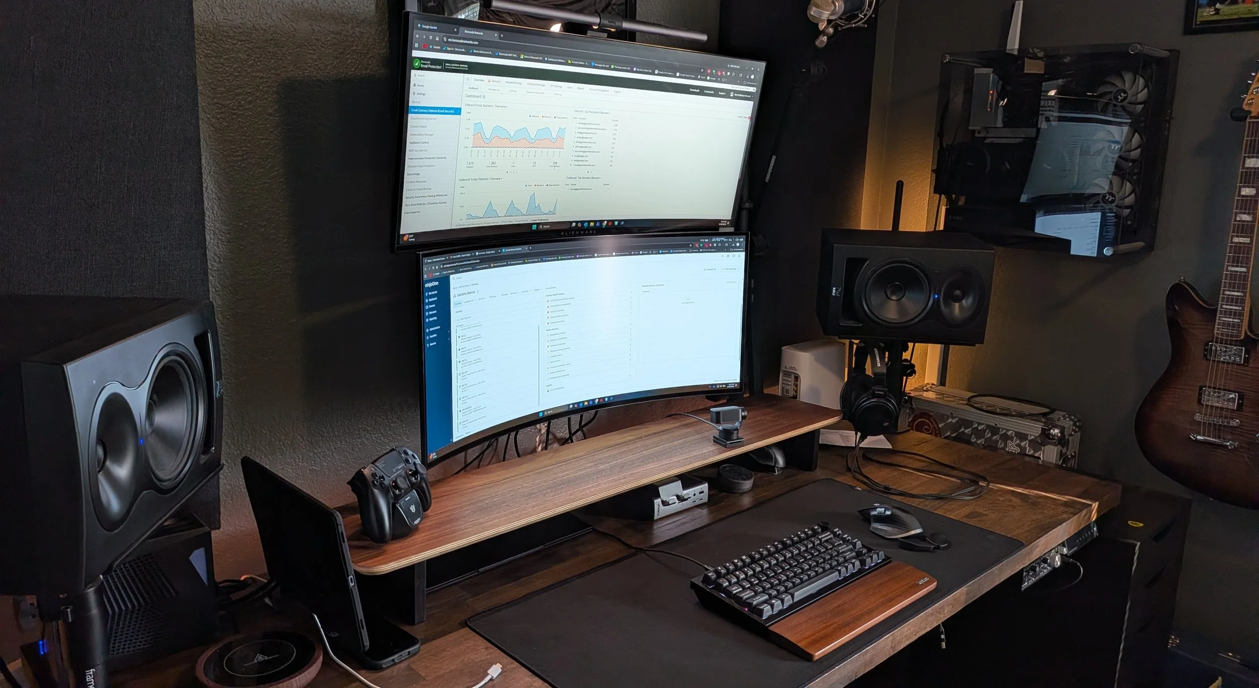 A computer desk setup with dual curved monitors displaying analytics and email, surrounded by speakers, a gaming controller, a phone, a keyboard with a wooden wrist rest, a mouse, headphones, and a desktop computer inside a transparent side case. A guitar is mounted on the wall to the right.