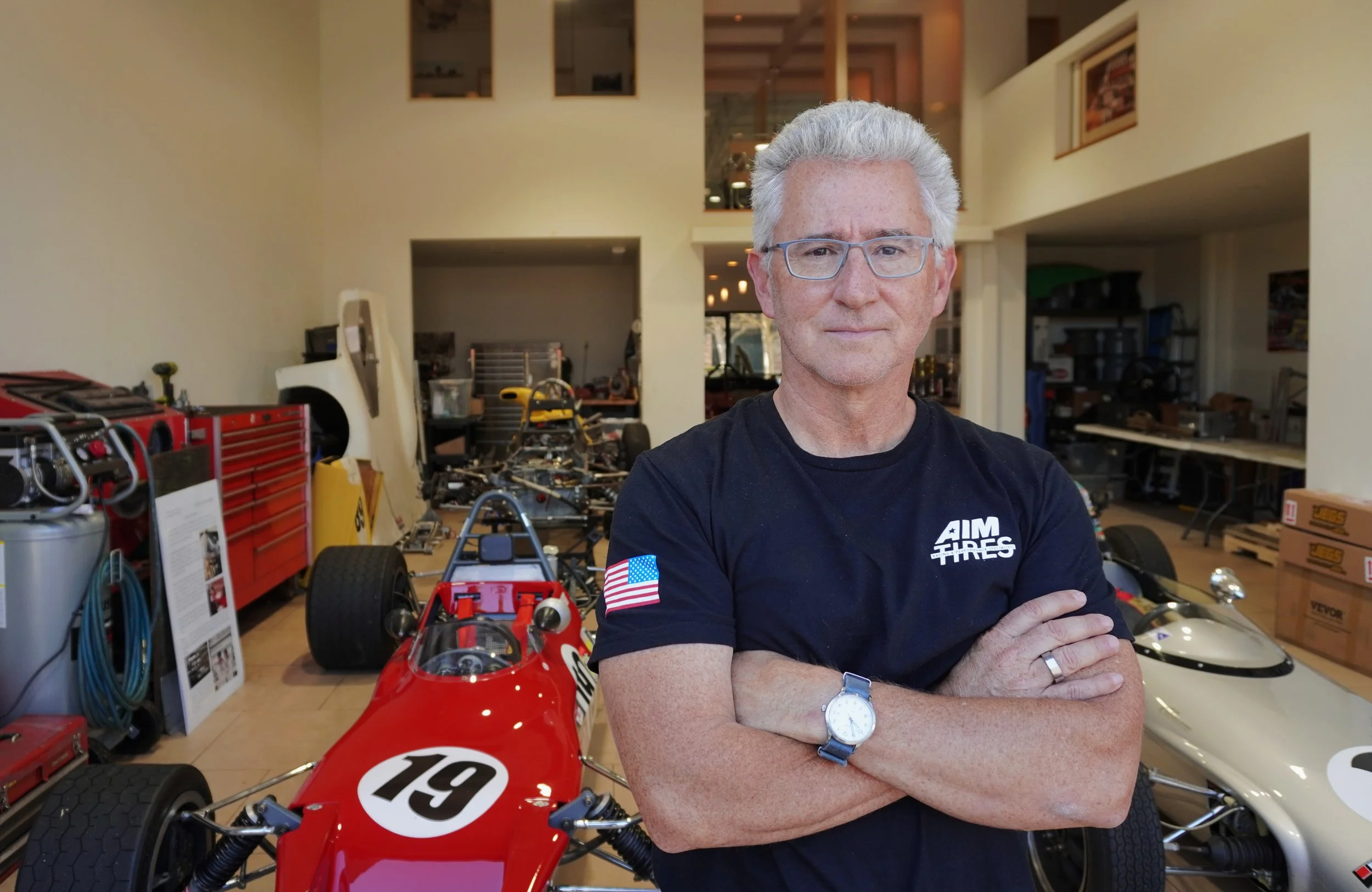 A man with gray hair and glasses crossing my arms, wearing a black t-shirt with an American flag patch on the sleeve, standing in front of a red race car with the number 19, in a workshop or garage filled with tools, car parts, and racing equipment.