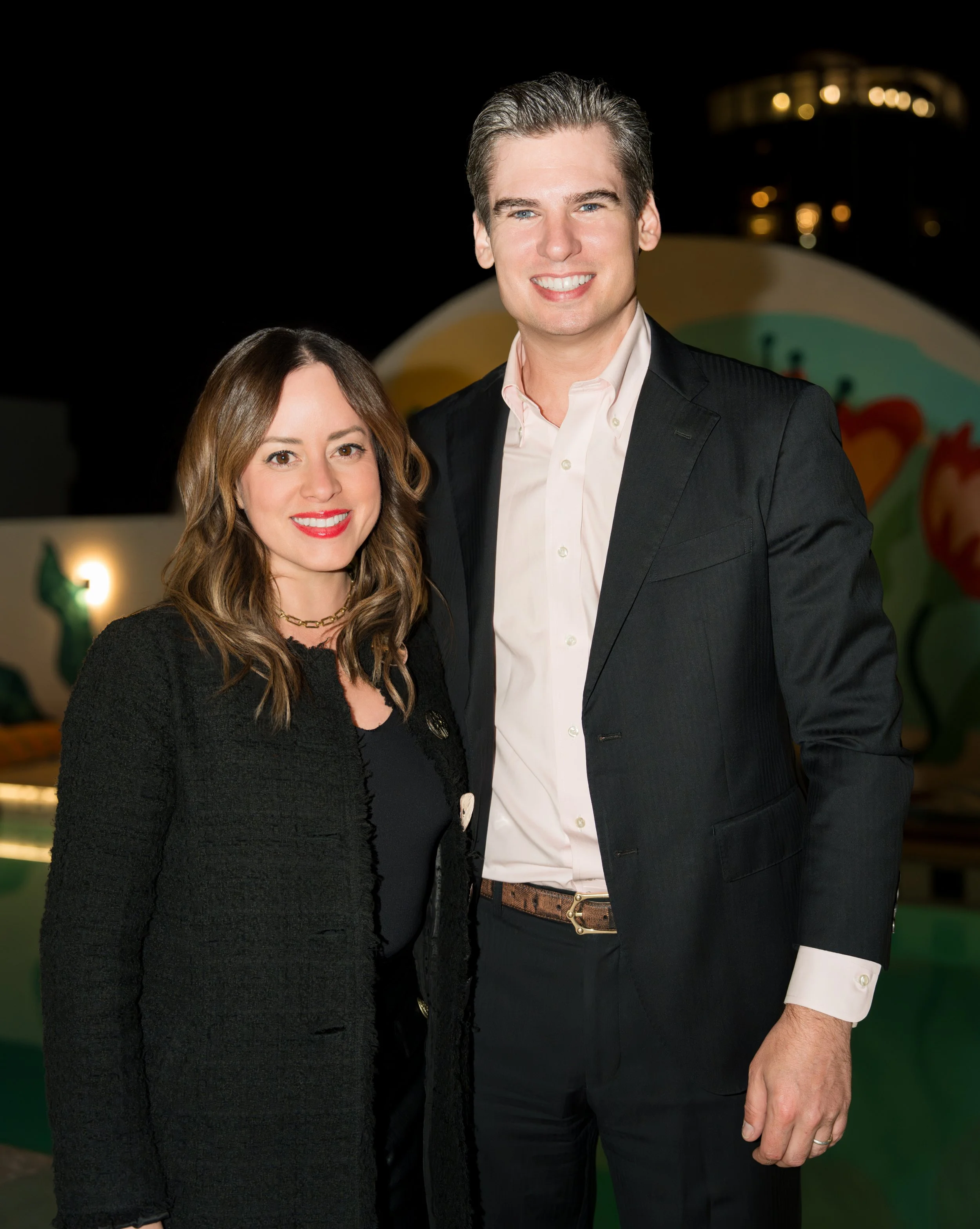 A smiling man and woman standing together outdoors at night, with colorful balloons and string lights in the background.