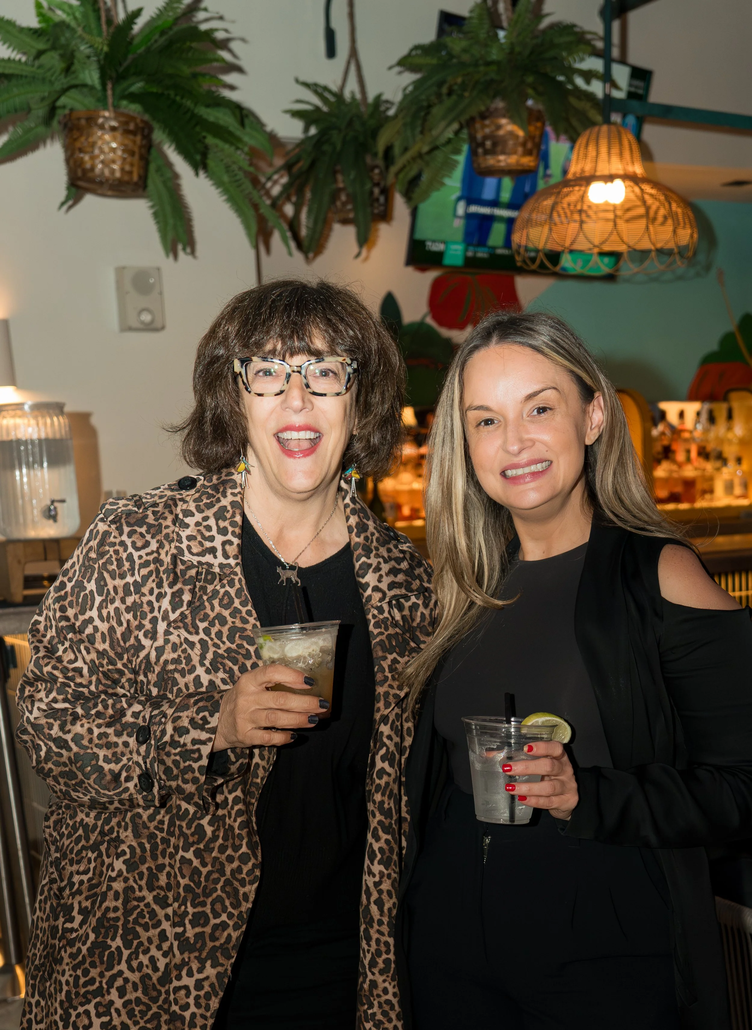 Two women standing together, smiling and holding drinks in a lively indoor setting with hanging plants, warm lighting, and a bar in the background.