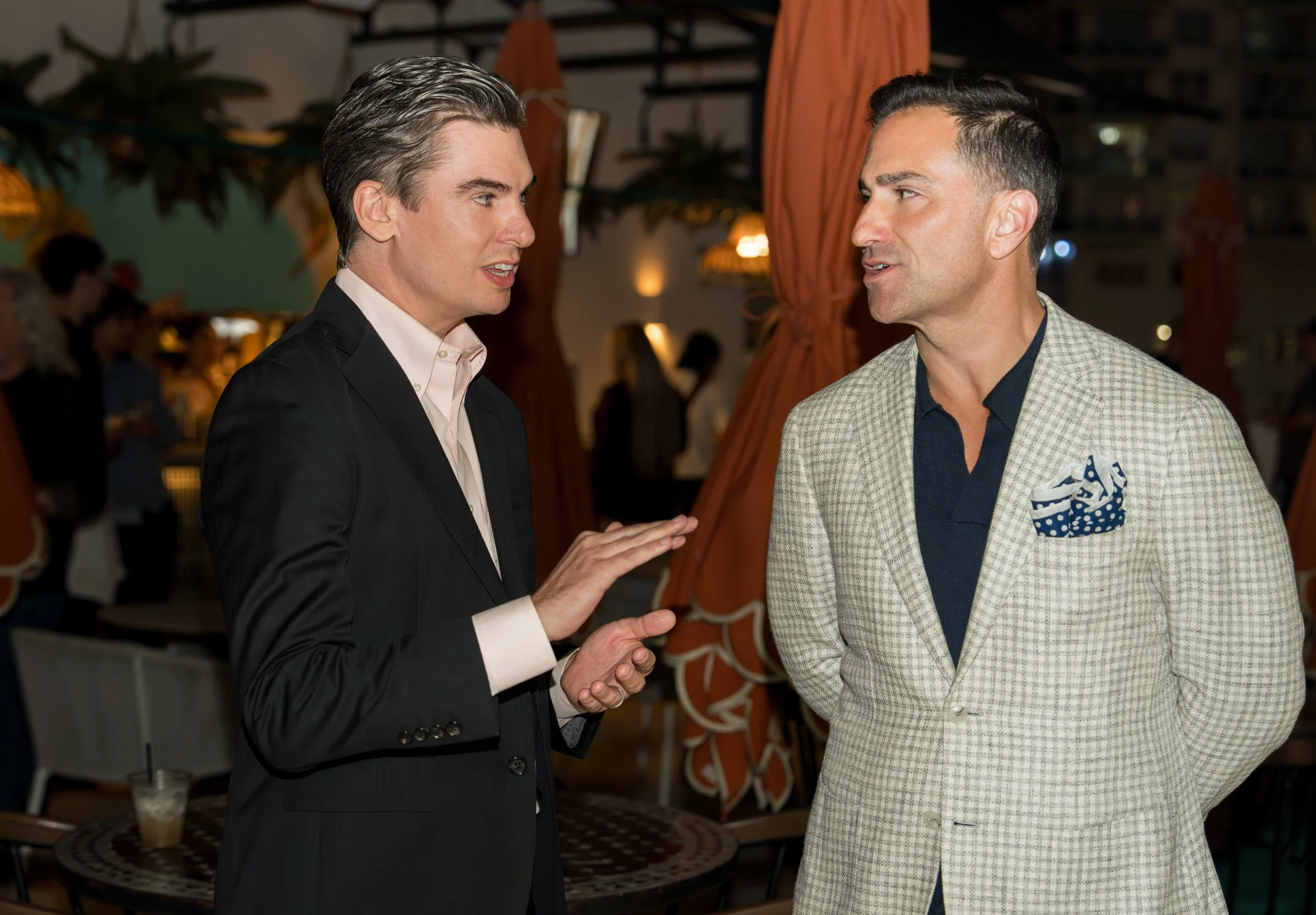 Two men having a conversation at an outdoor event or restaurant at night, dressed in business casual attire. One man is speaking and gesturing with his hands, while the other listens attentively.