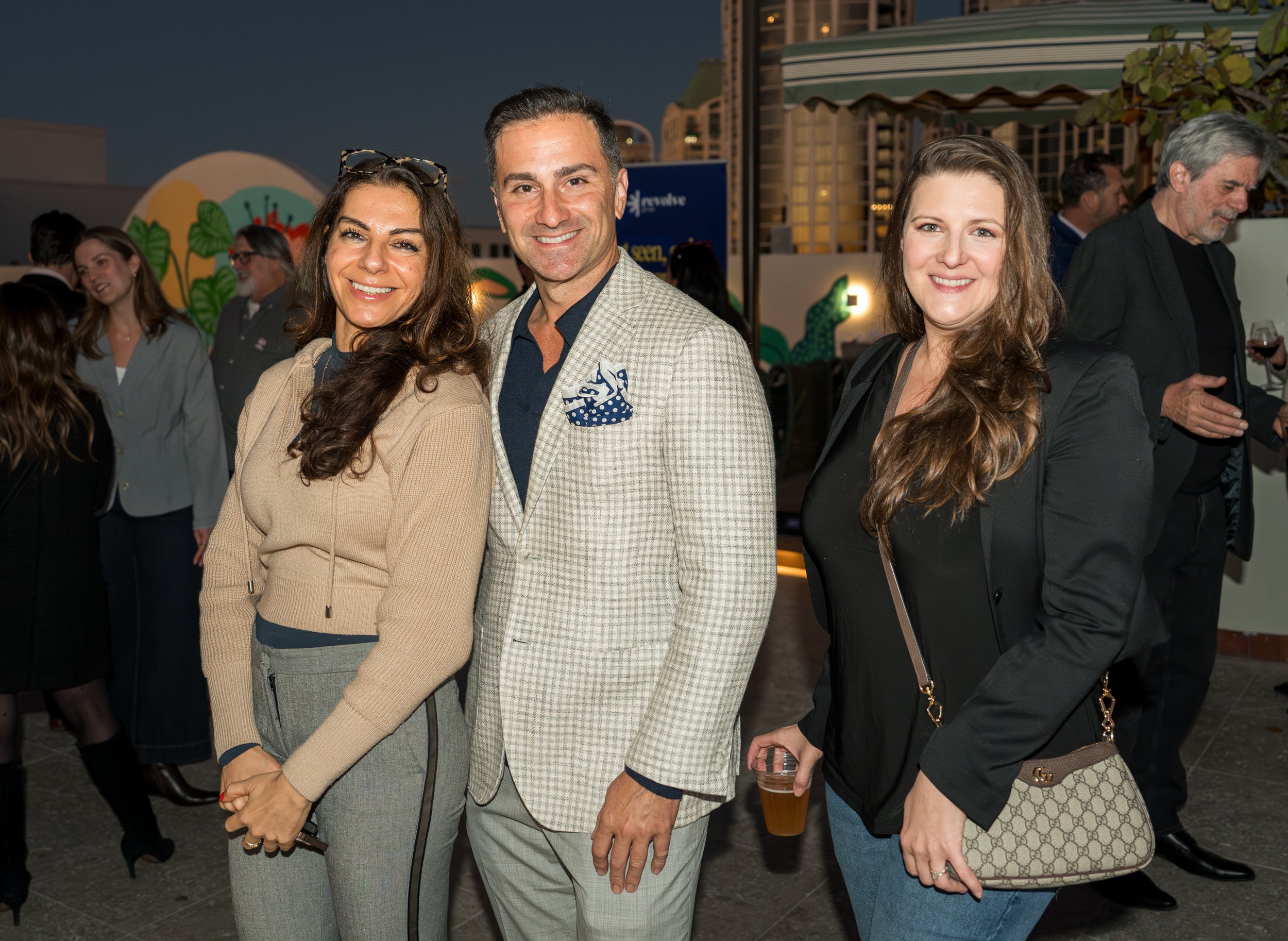Three people standing together at a social outdoor event during evening, smiling at the camera, with other guests in the background.