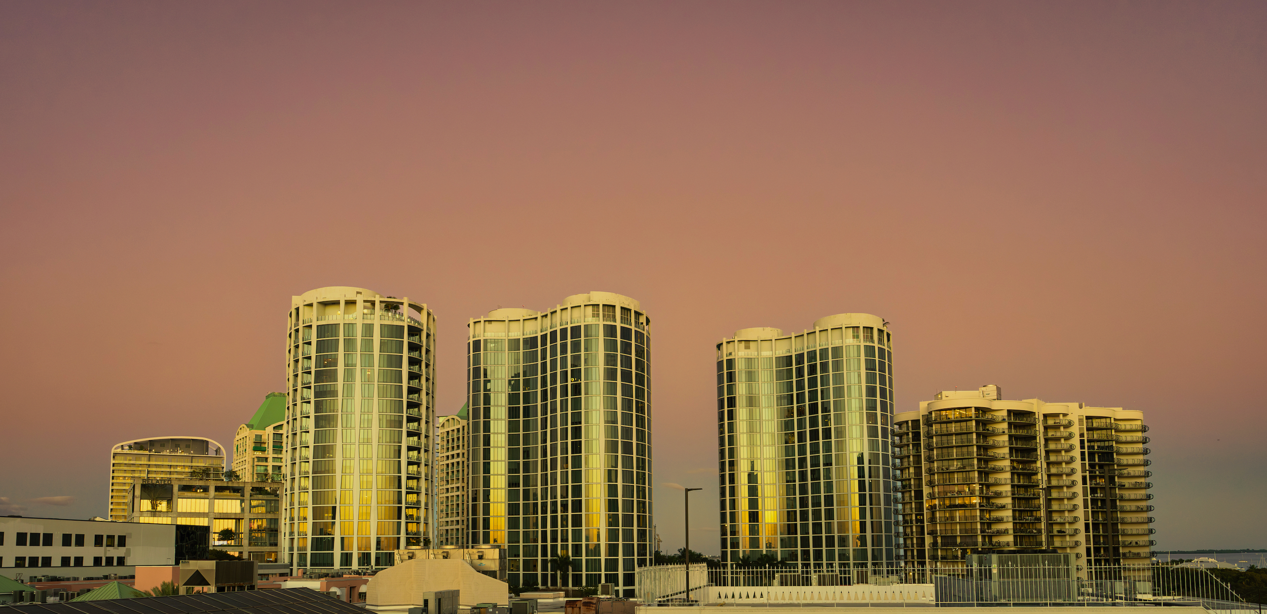 Skyline of modern high-rise buildings with glass windows, during sunset or sunrise, reflecting warm colors.