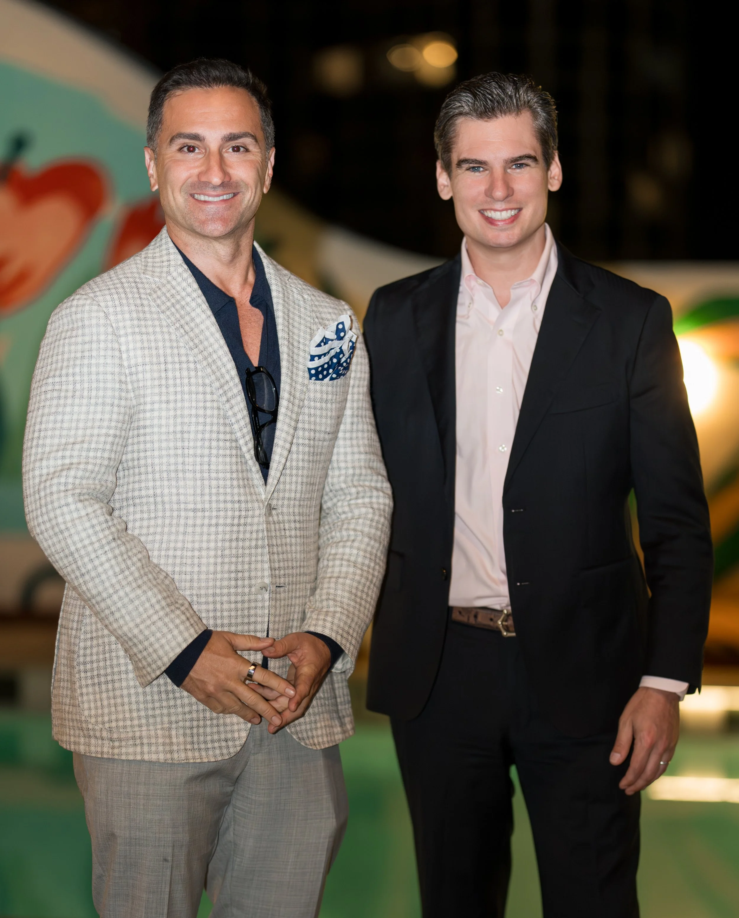 Two men in suits standing side by side smiling at the camera, with blurred colorful background.