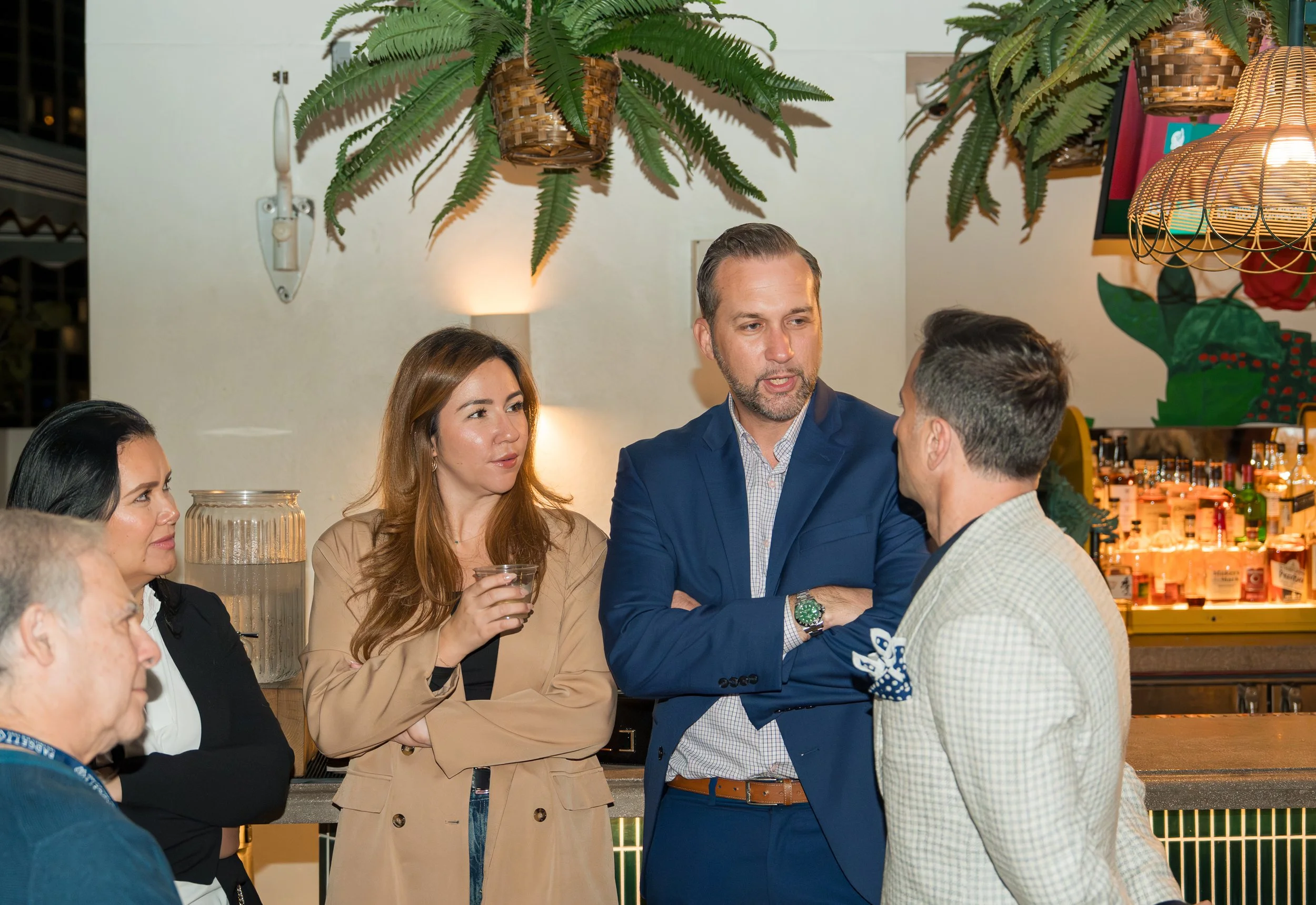 Group of five people having a conversation at a social gathering in a bar or restaurant, with hanging plants and decorative lighting in the background.