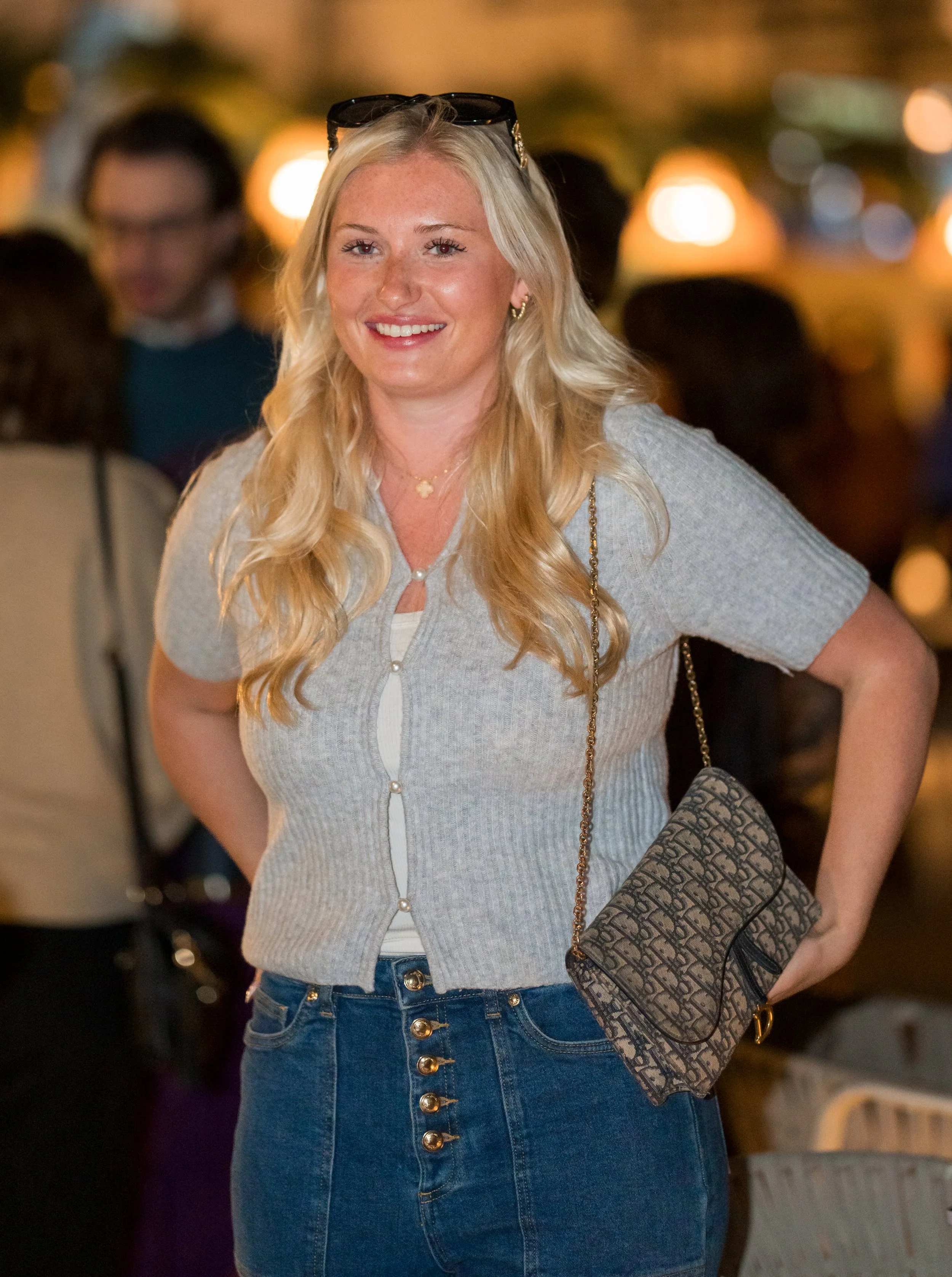 A blonde woman smiling outdoors at night, wearing a gray cardigan, blue jeans, sunglasses on her head, and carrying a patterned purse.