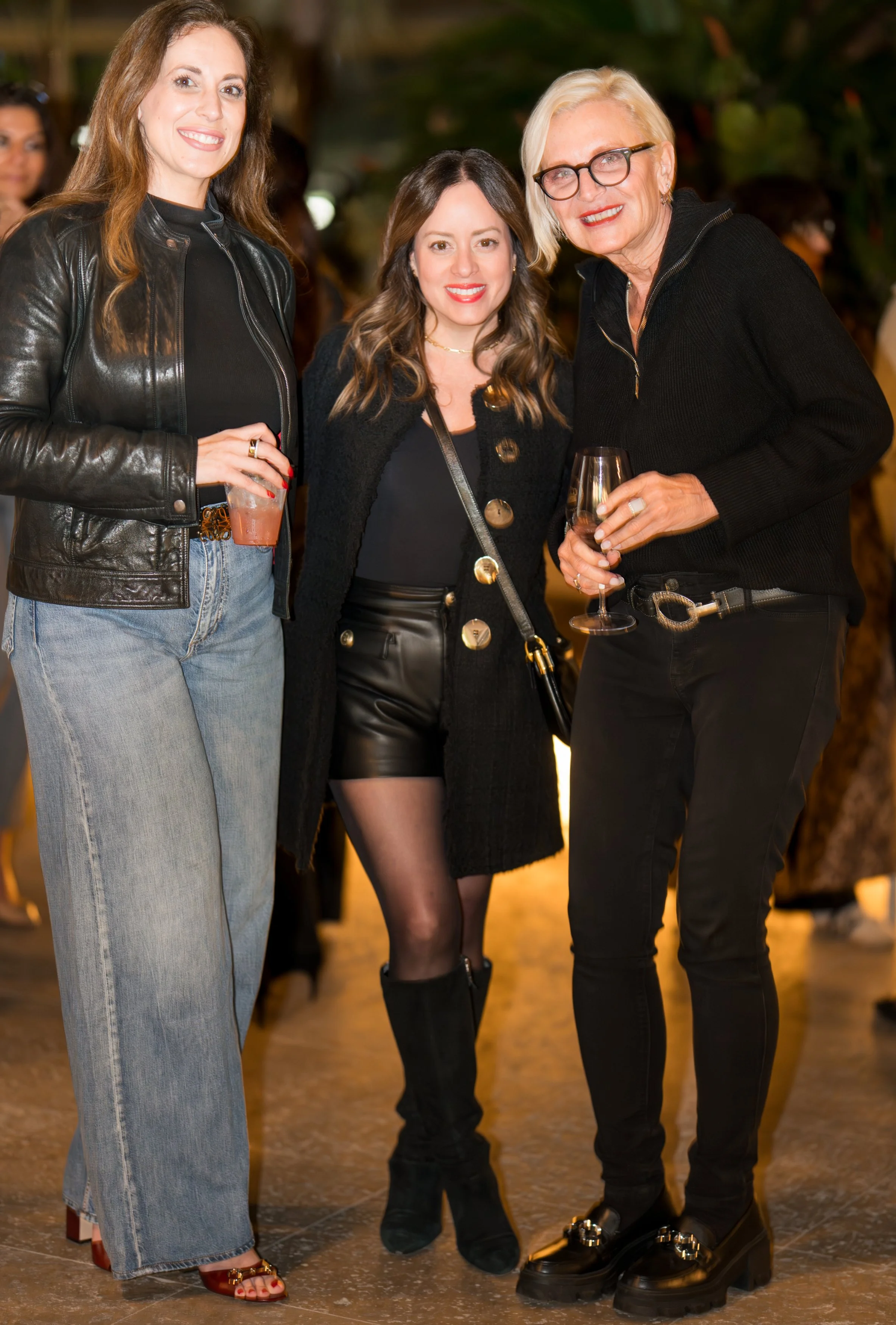 Three women at a social event holding drinks, smiling for the camera. The woman on the left is wearing a black leather jacket and jeans, the woman in the middle is in a black top with a black coat and leather shorts, and the woman on the right is wea