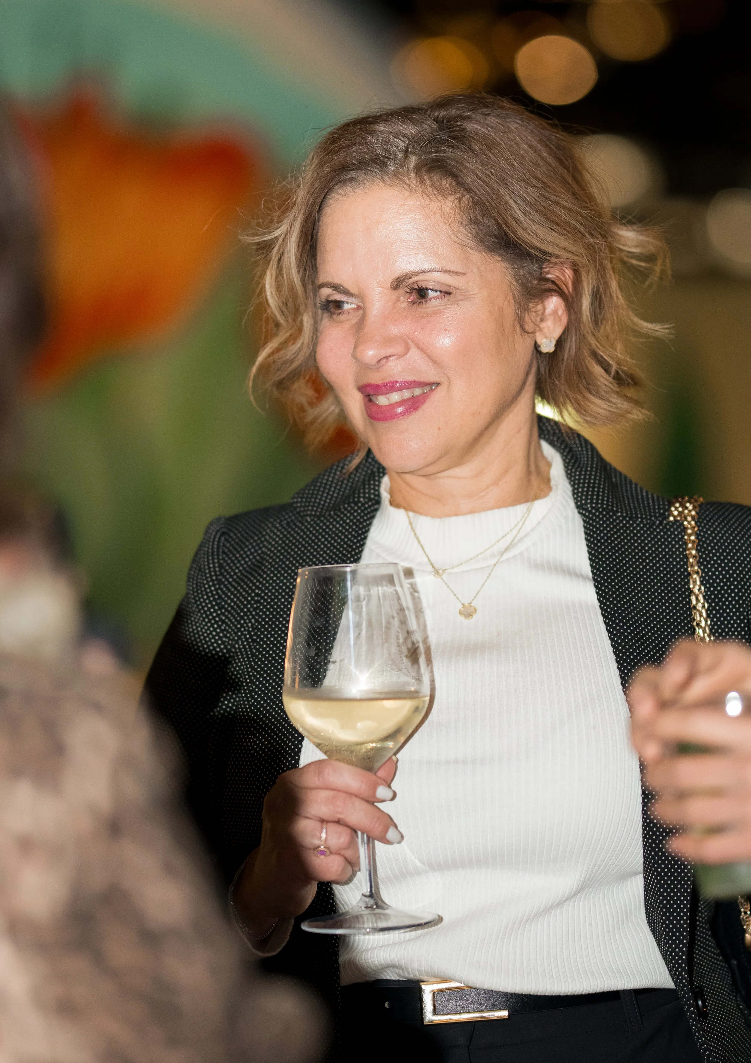 A woman with short, wavy brown hair, wearing a white top and black blazer with white polka dots, holding a glass of white wine at a social event, smiling and looking to her right.