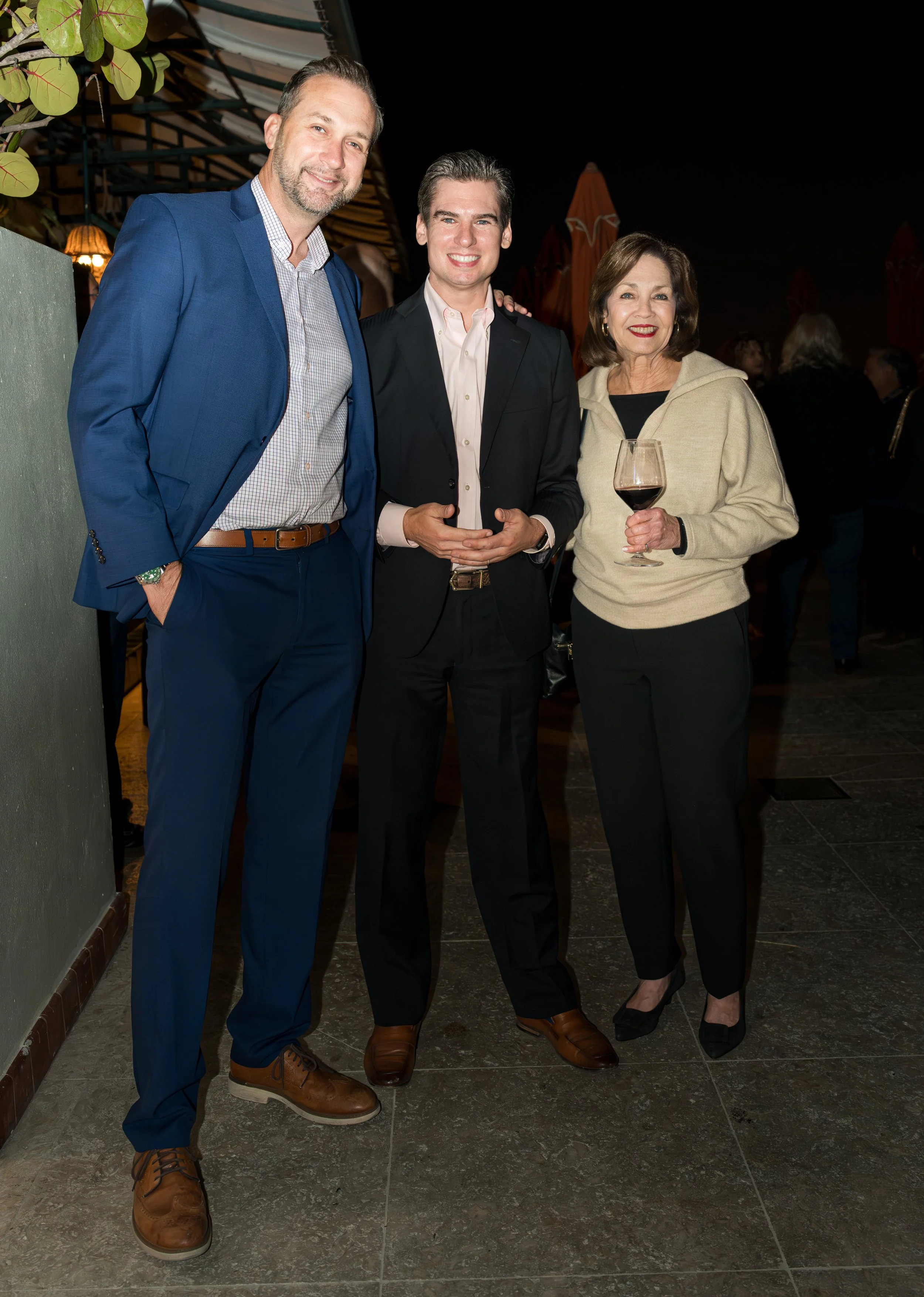 Three people at a social event, smiling at the camera. The man on the left is wearing a blue suit and brown shoes, the man in the middle is dressed in a black suit and holding his hands together, and the woman on the right is holding a glass of red w