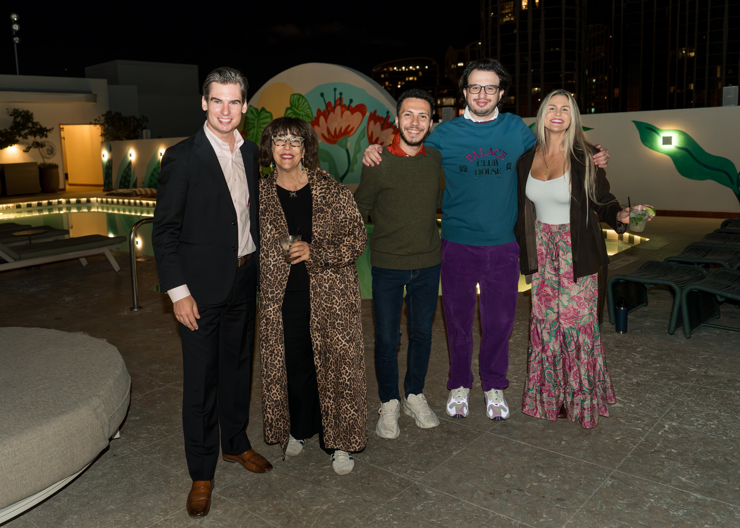 A group of six people standing together at night by a poolside with city buildings in the background, smiling and posing for the photo.