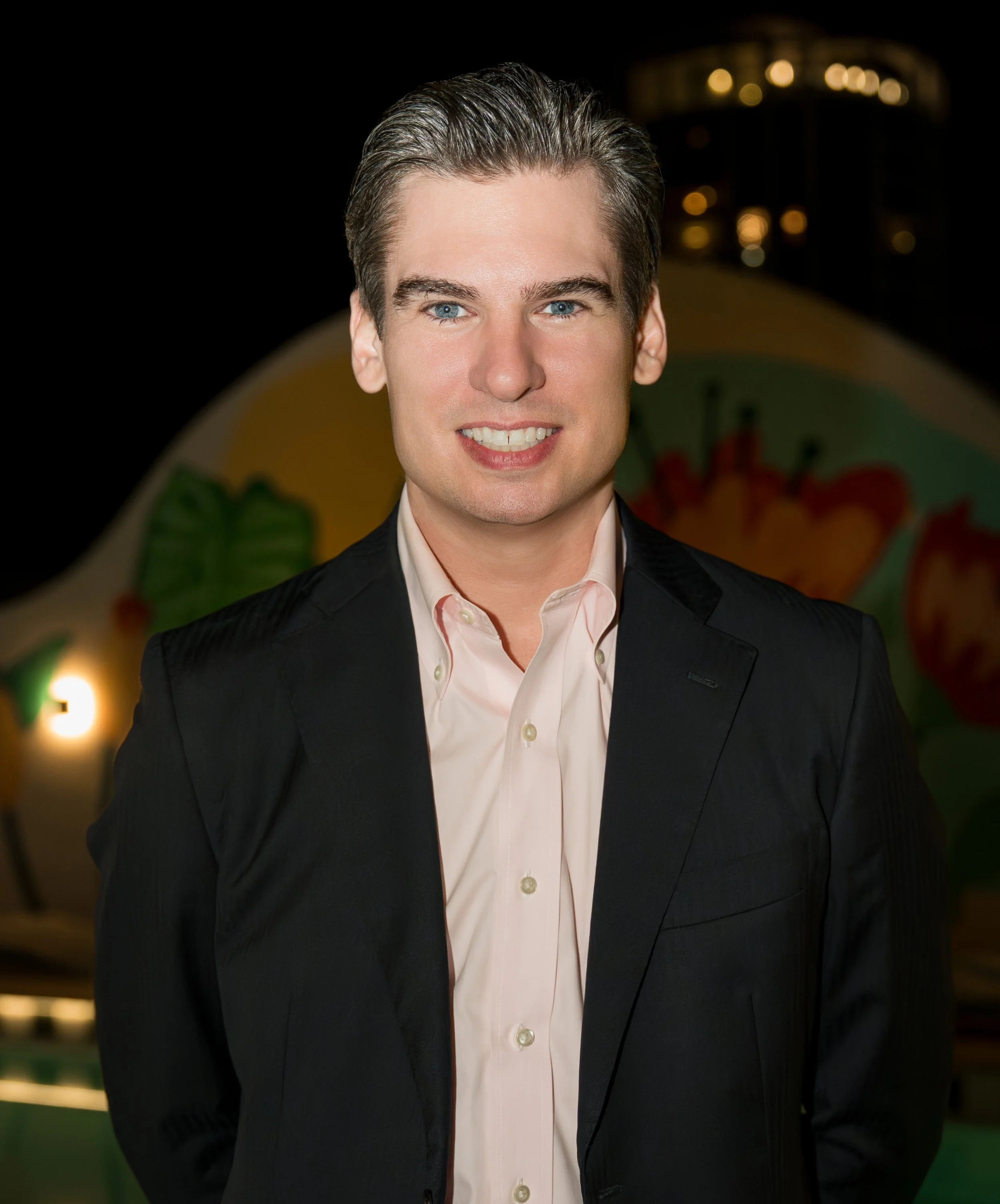 Portrait of a young man with slicked-back hair, wearing a light pink dress shirt and dark blazer, smiling at the camera at night with a colorful carnival ride in the background.